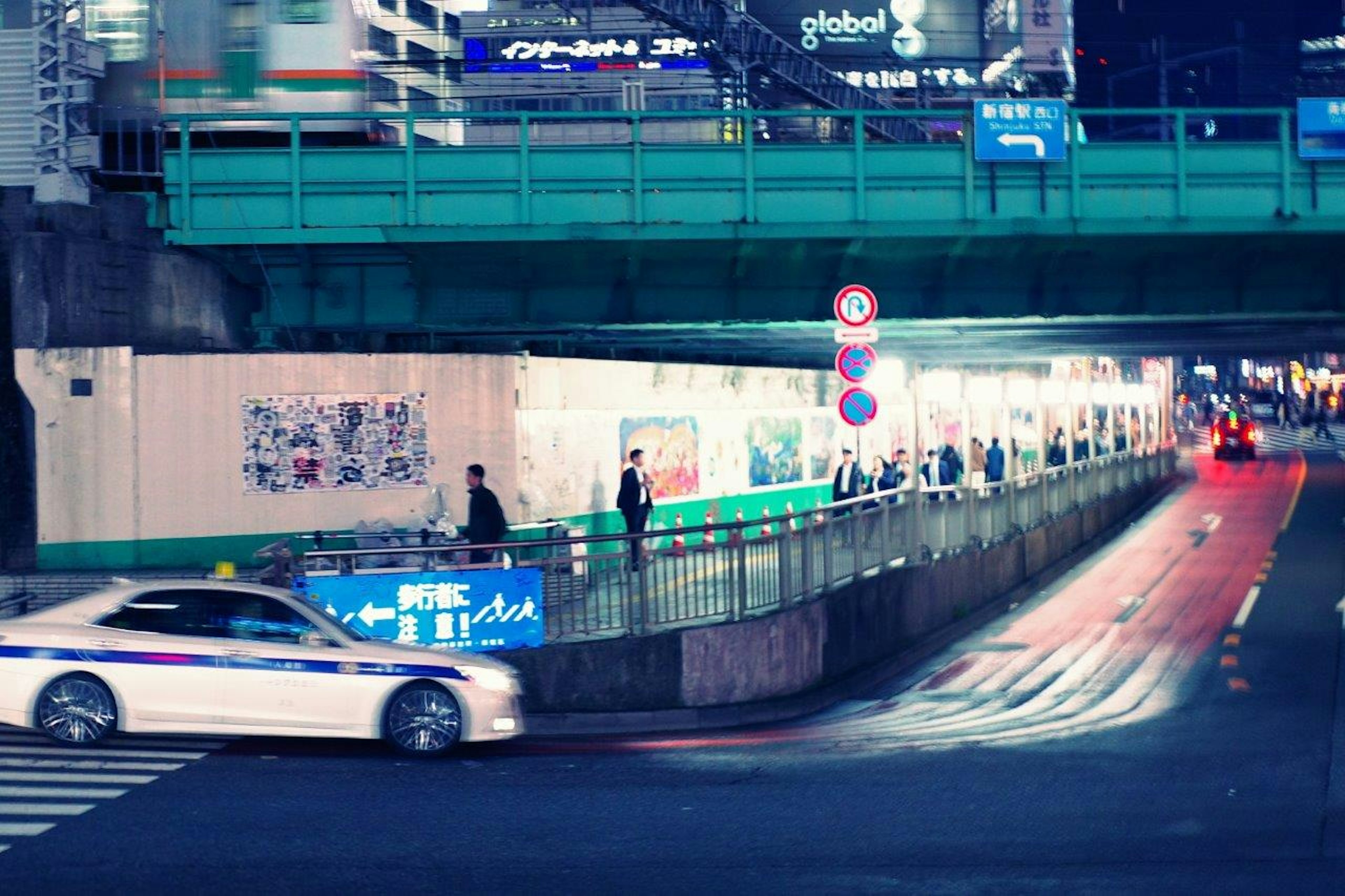 Scène urbaine nocturne avec une voiture de police blanche circulant sous un pont bleu et des piétons sur le trottoir