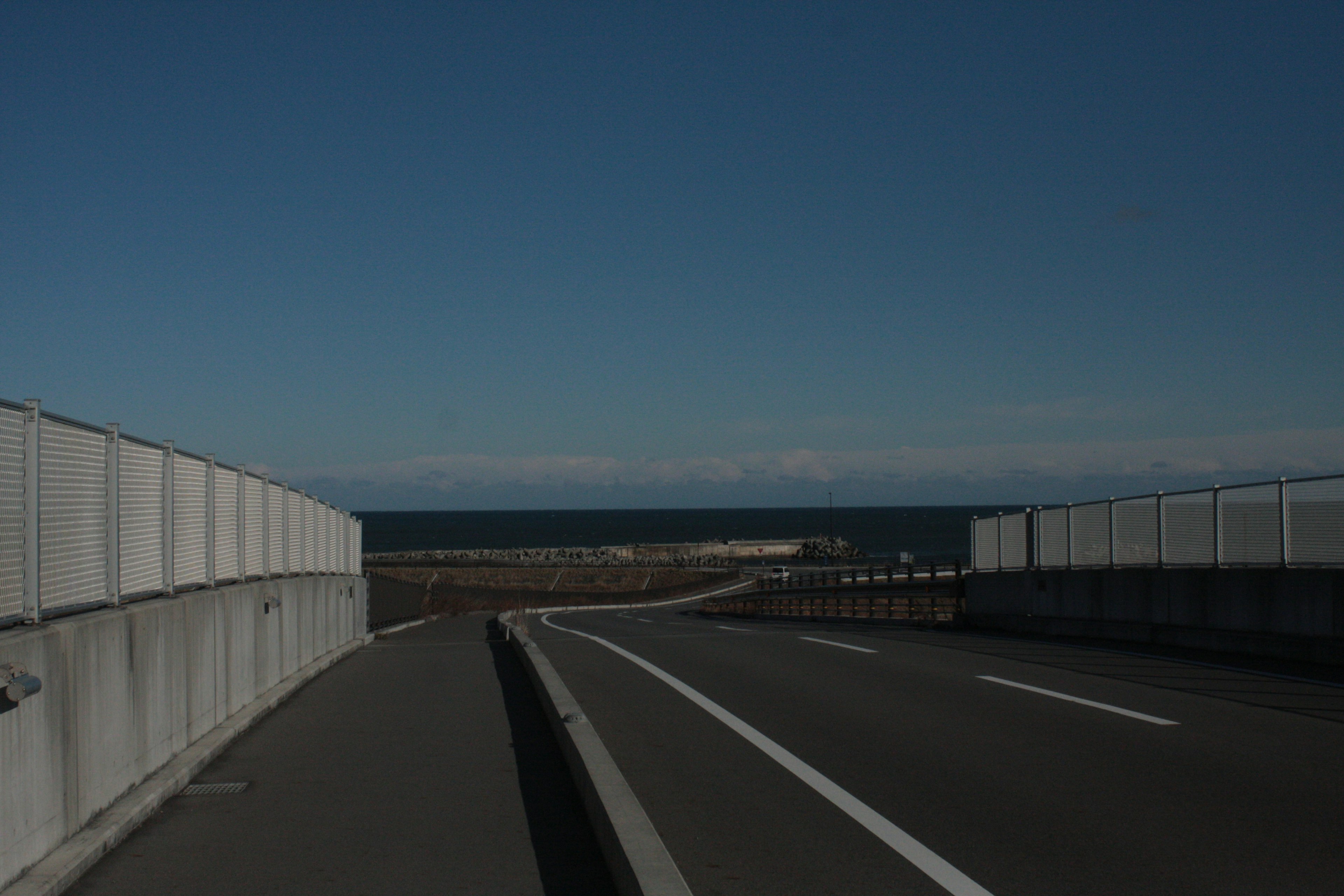 Curving road leading to the sea under a blue sky with white fence