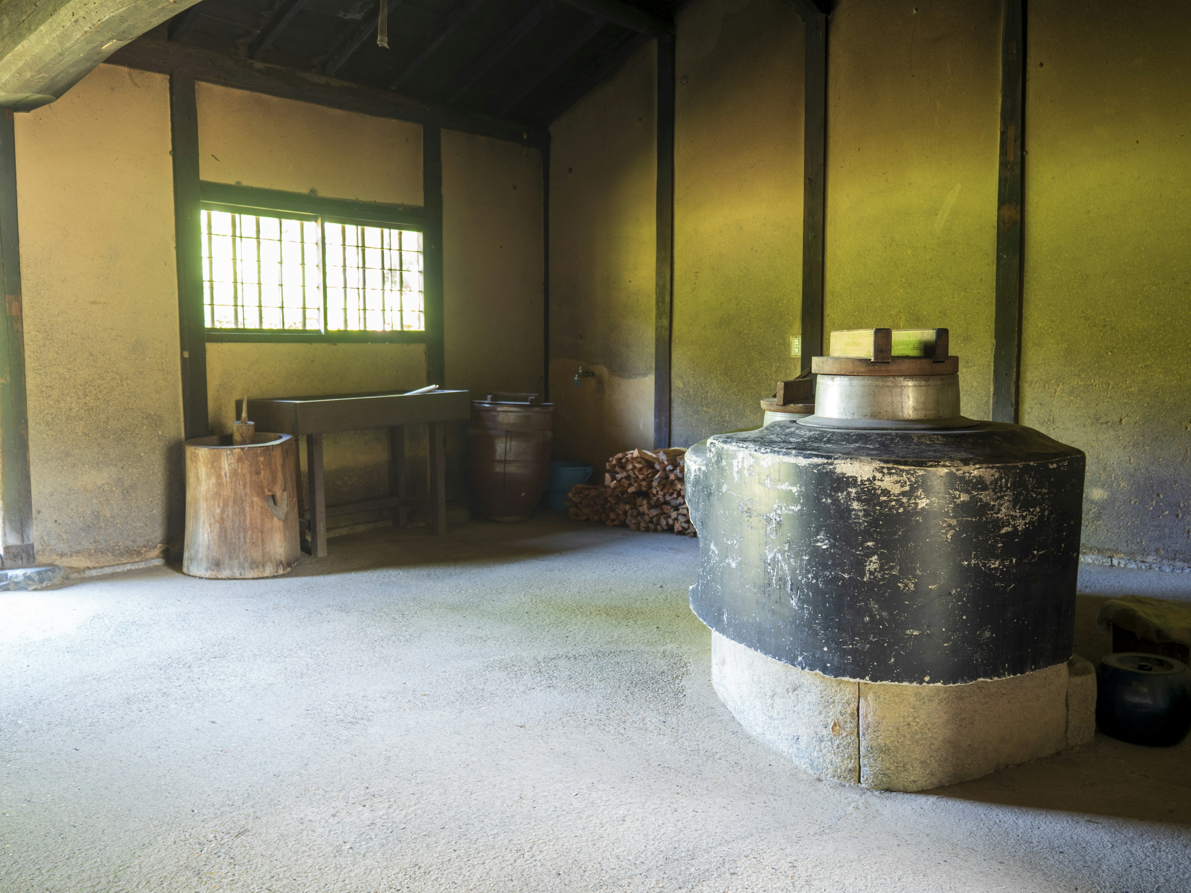 Interior view of an old Japanese building with concrete floor and greenish walls featuring a large black stove on the right and a wooden table on the left