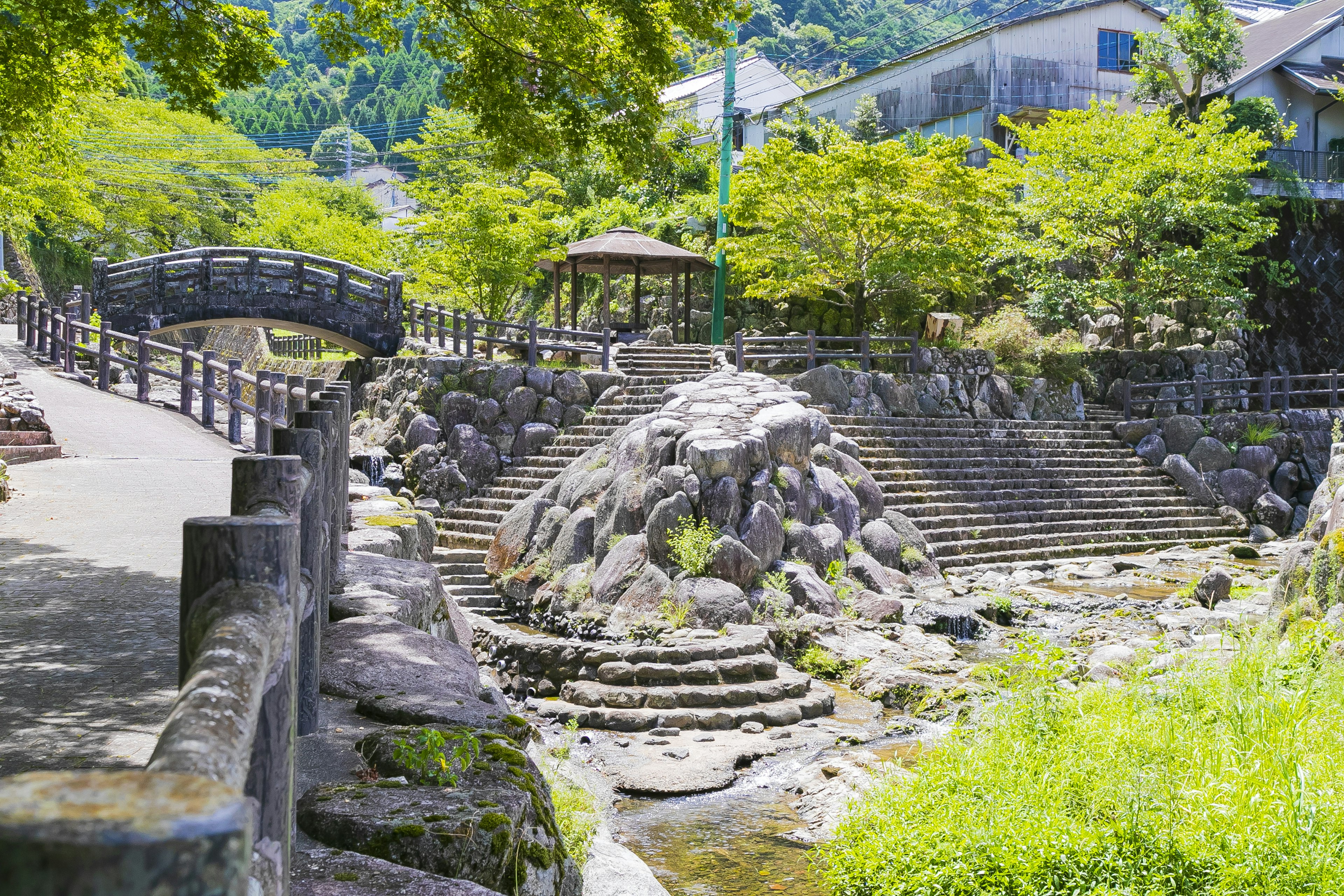 Vista escénica con escalones de piedra y un arroyo rodeado de vegetación puente tradicional y gazebo visibles