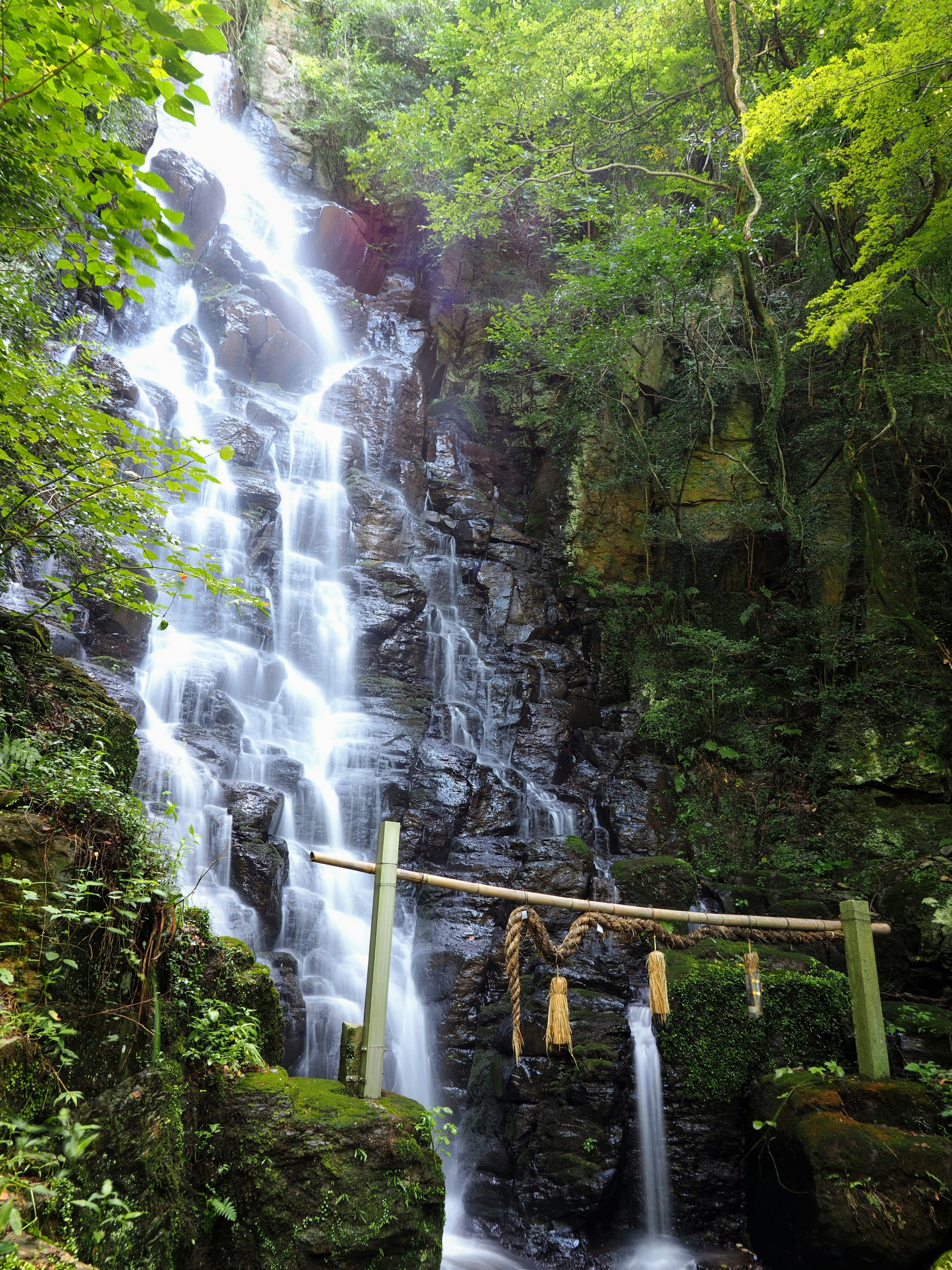 Una cascada que cae sobre un terreno rocoso con abundante vegetación y un puente de madera sagrado