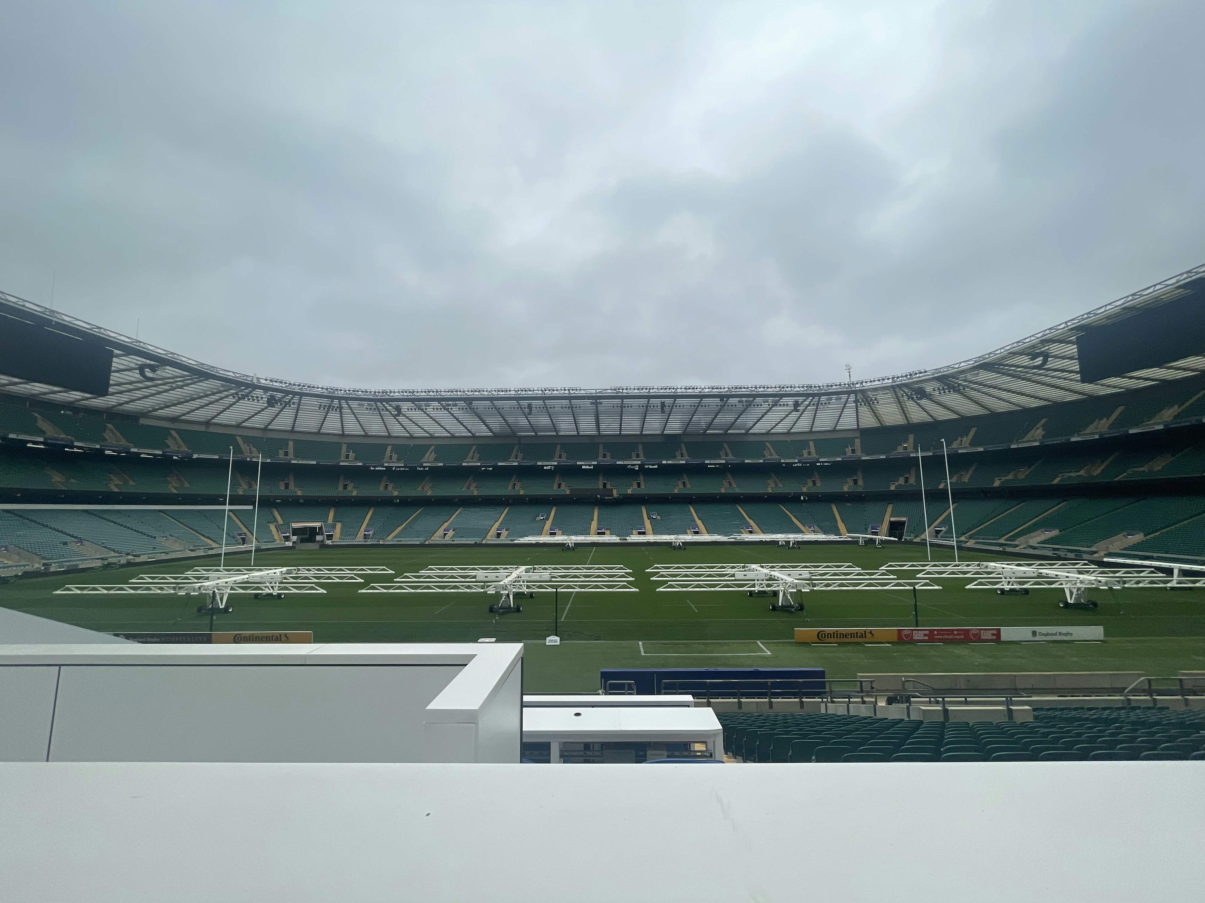 Interior stadion dengan rumput hijau, langit mendung, area tempat duduk terlihat