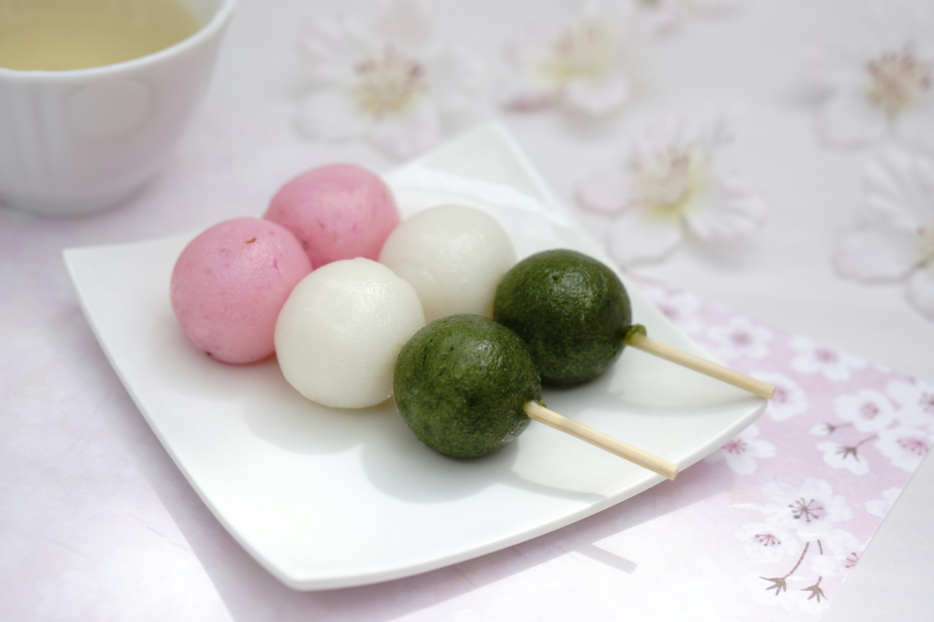 Tre spiedini di dango rosa, bianchi e verdi su un piatto bianco con uno sfondo floreale