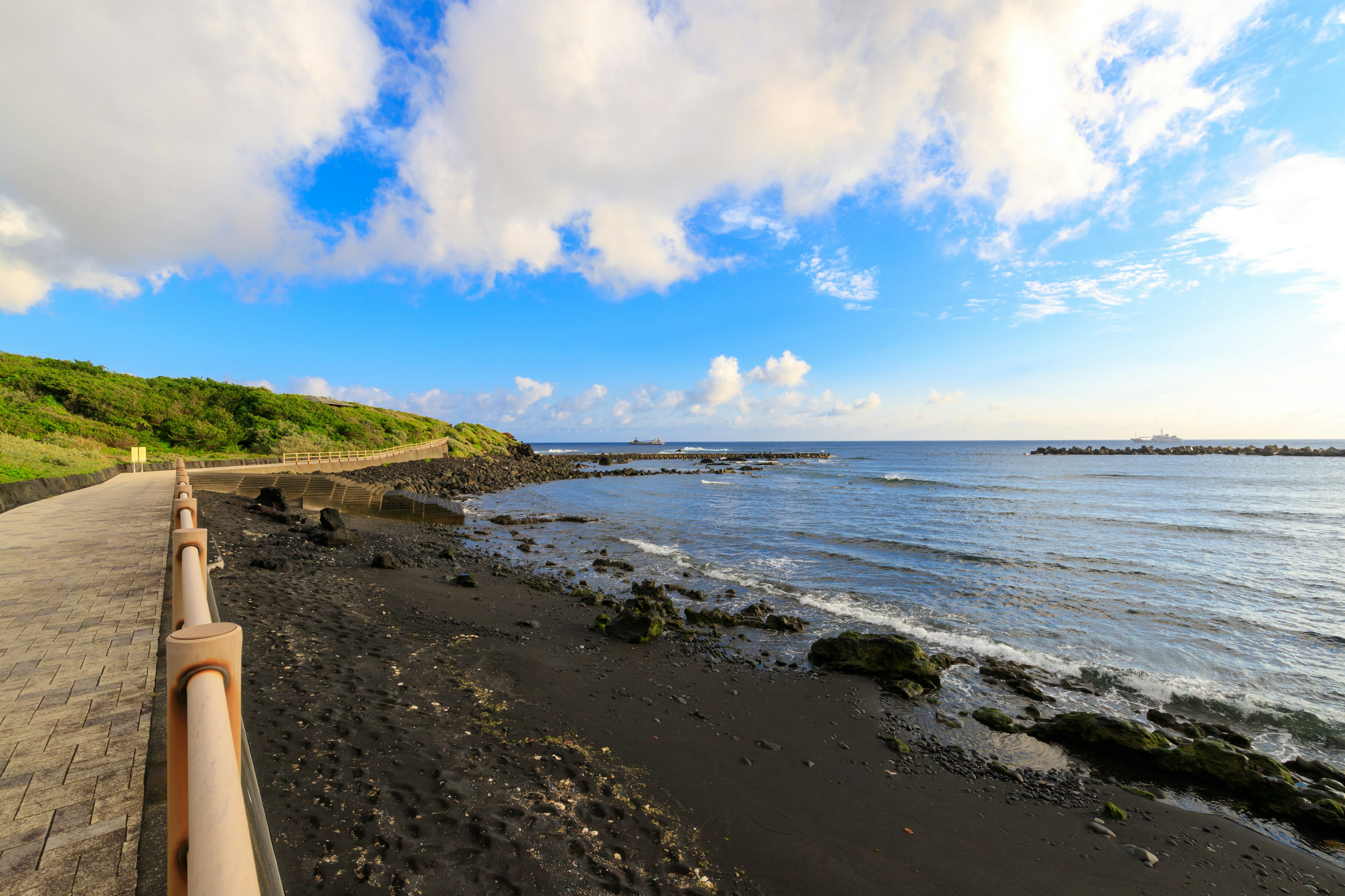 Küstenweg mit schwarzem Sand und Wellen unter blauem Himmel