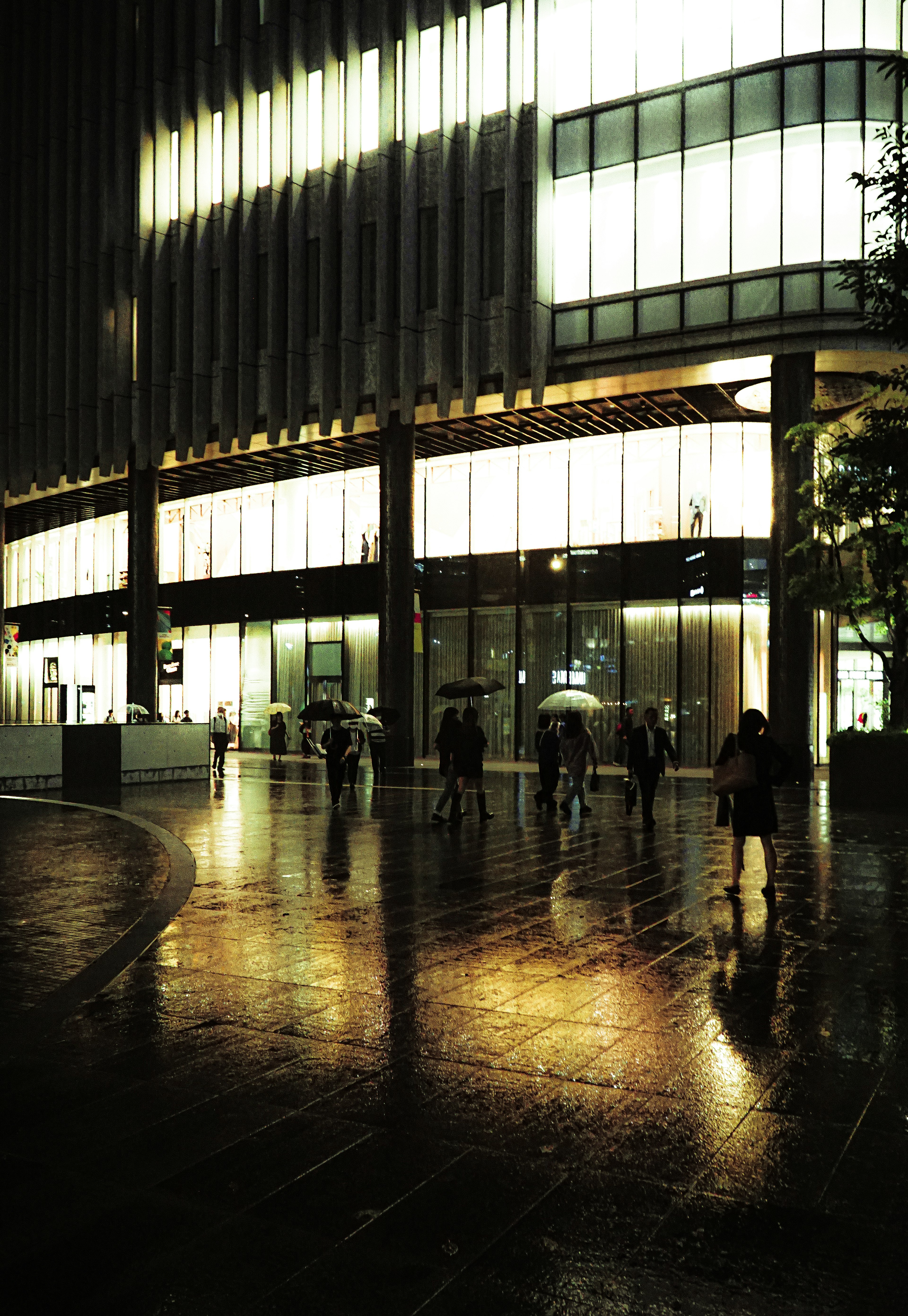 Bâtiment moderne la nuit avec des reflets sur le pavé mouillé et des personnes marchant