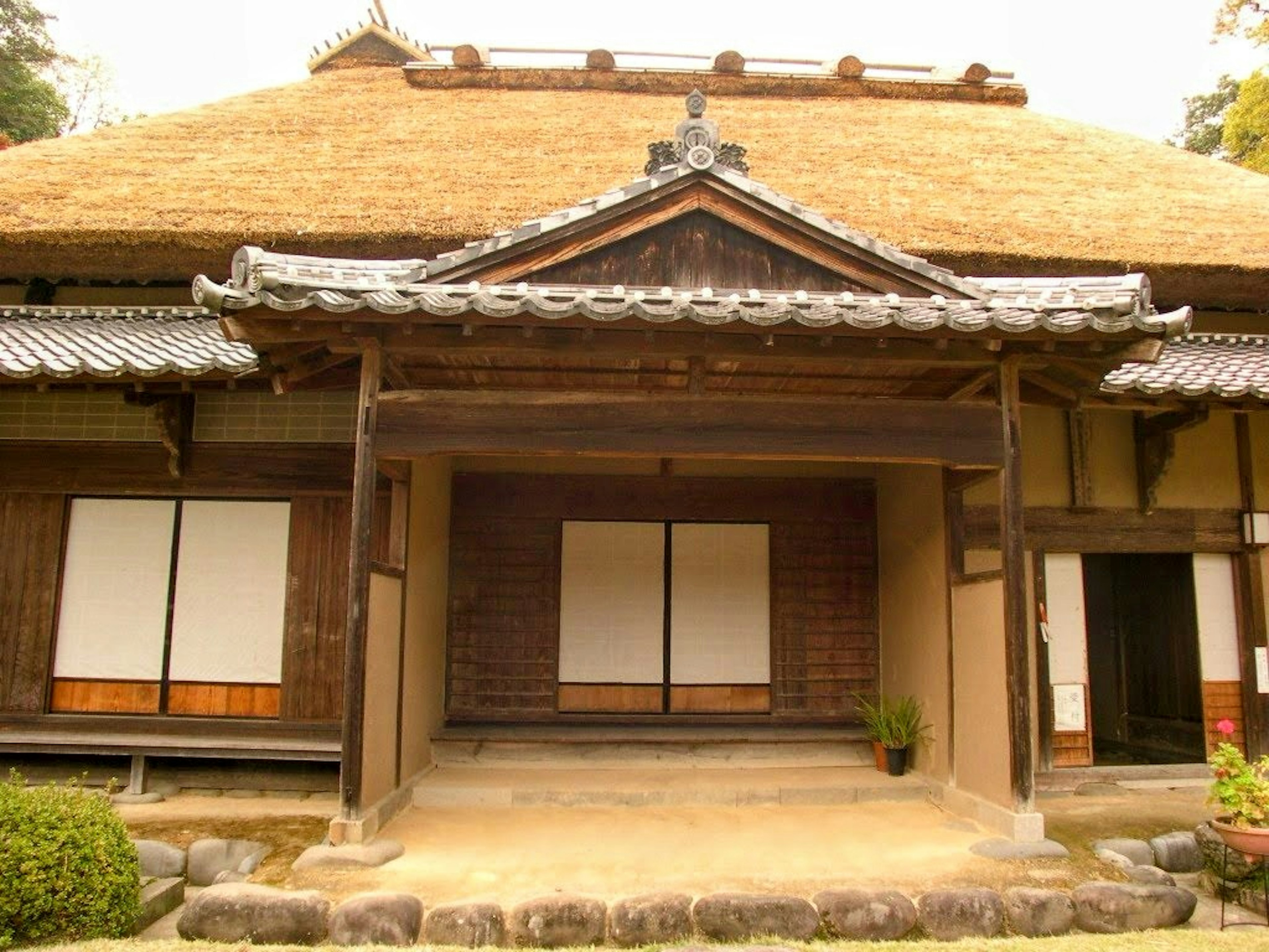 Traditional Japanese house exterior with thatched roof and wooden structure surrounded by greenery