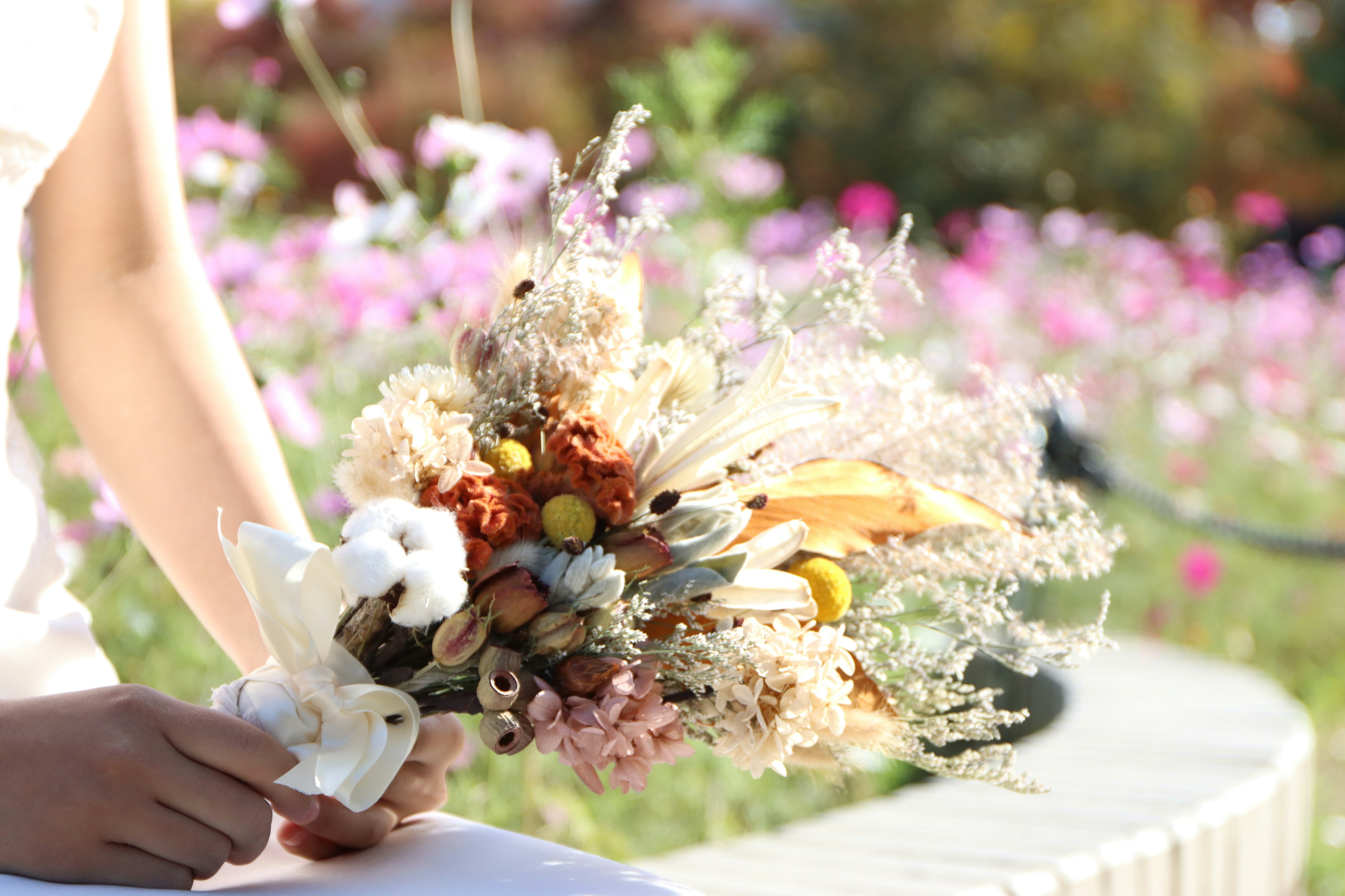 Eine Hand einer Frau, die einen bunten Trockenblumenstrauß hält, mit blühenden Blumen im Hintergrund