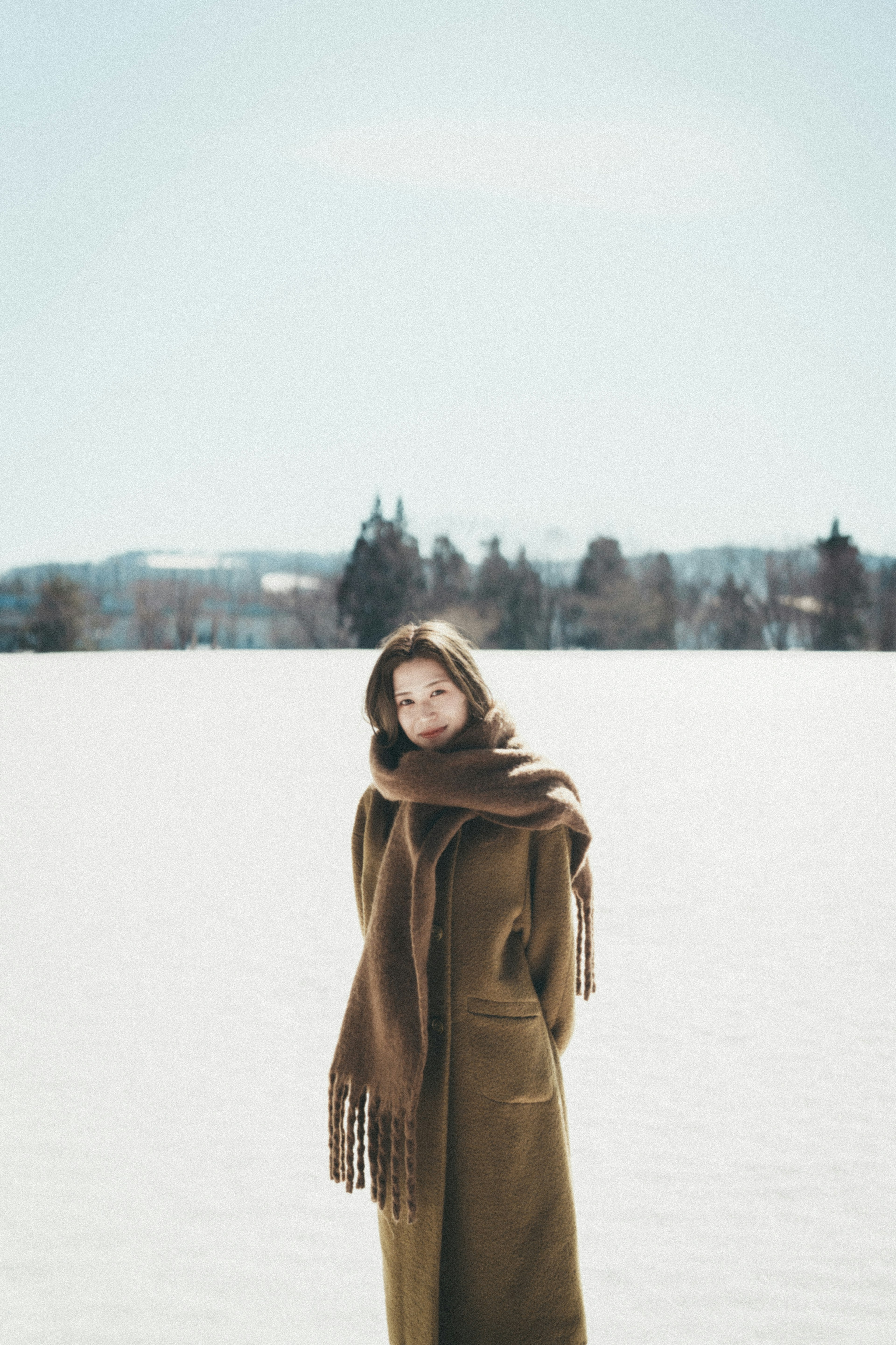 Eine Frau steht in einer schneebedeckten Landschaft mit einem hellbraunen Mantel und Schal