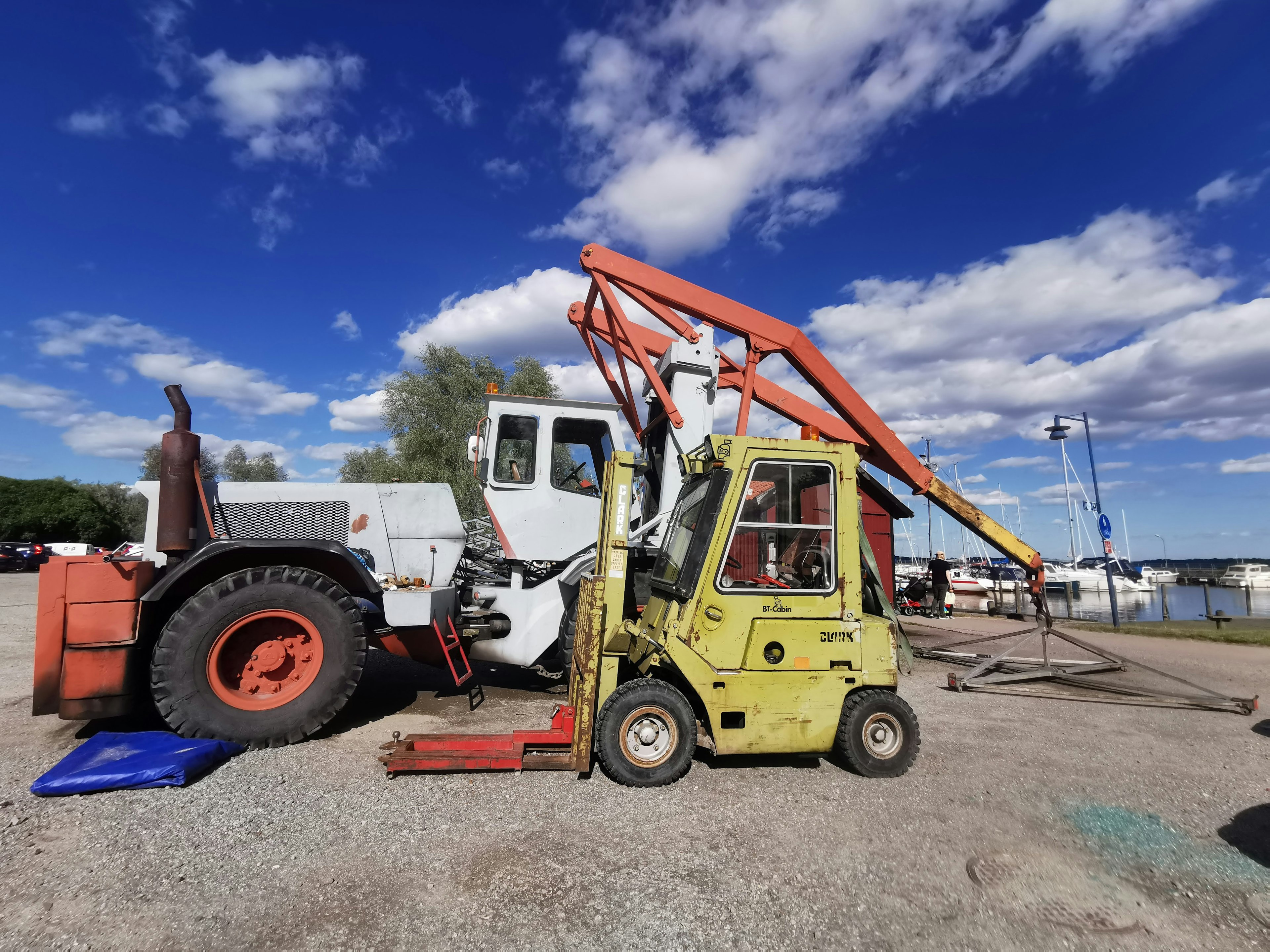 Sebuah forklift kuning dan traktor putih berdampingan dengan langit biru dan awan putih di latar belakang