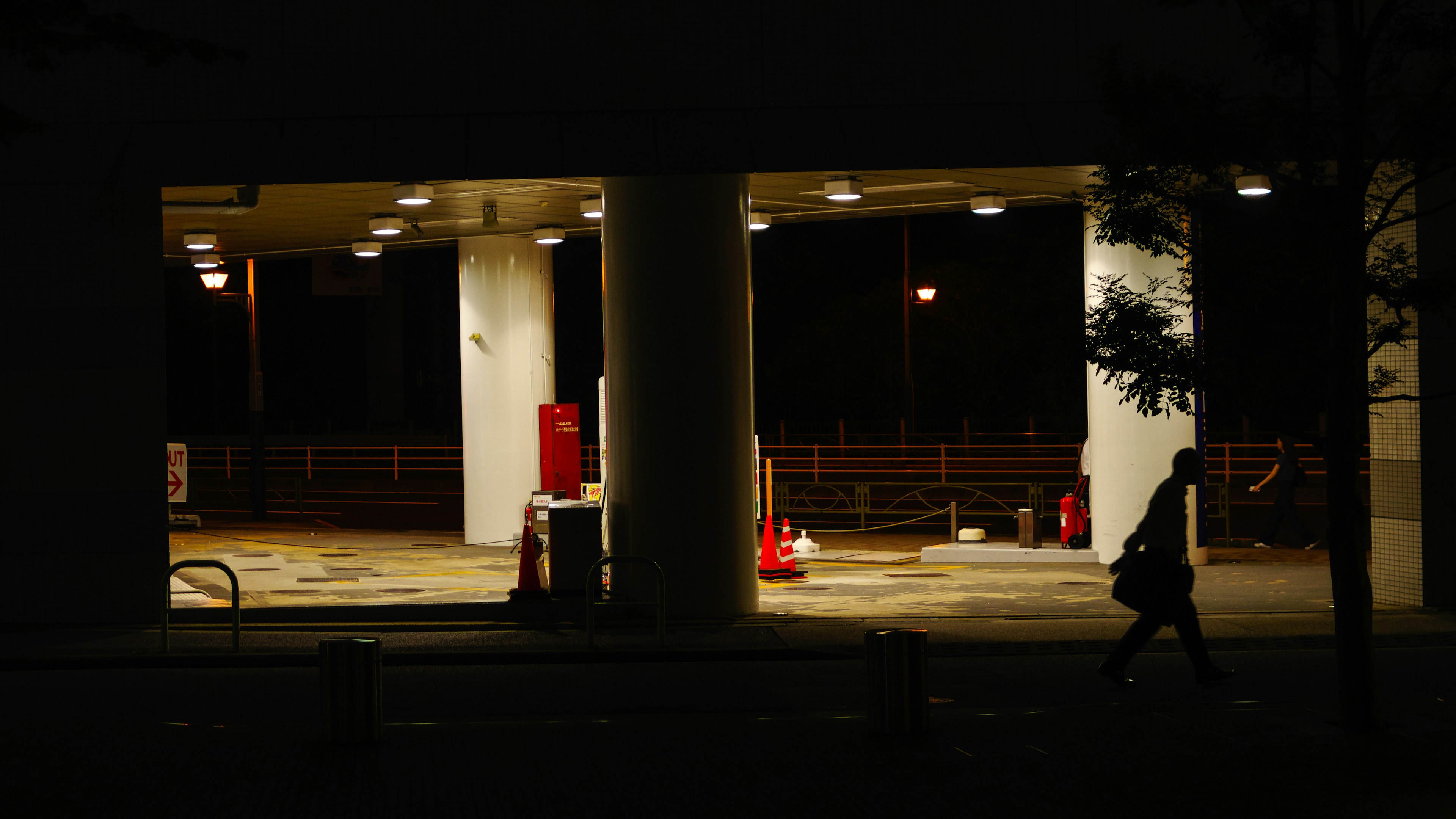 Silhouette einer Person, die nachts an einer Tankstelle mit roten Feuerlöschern vorbeigeht