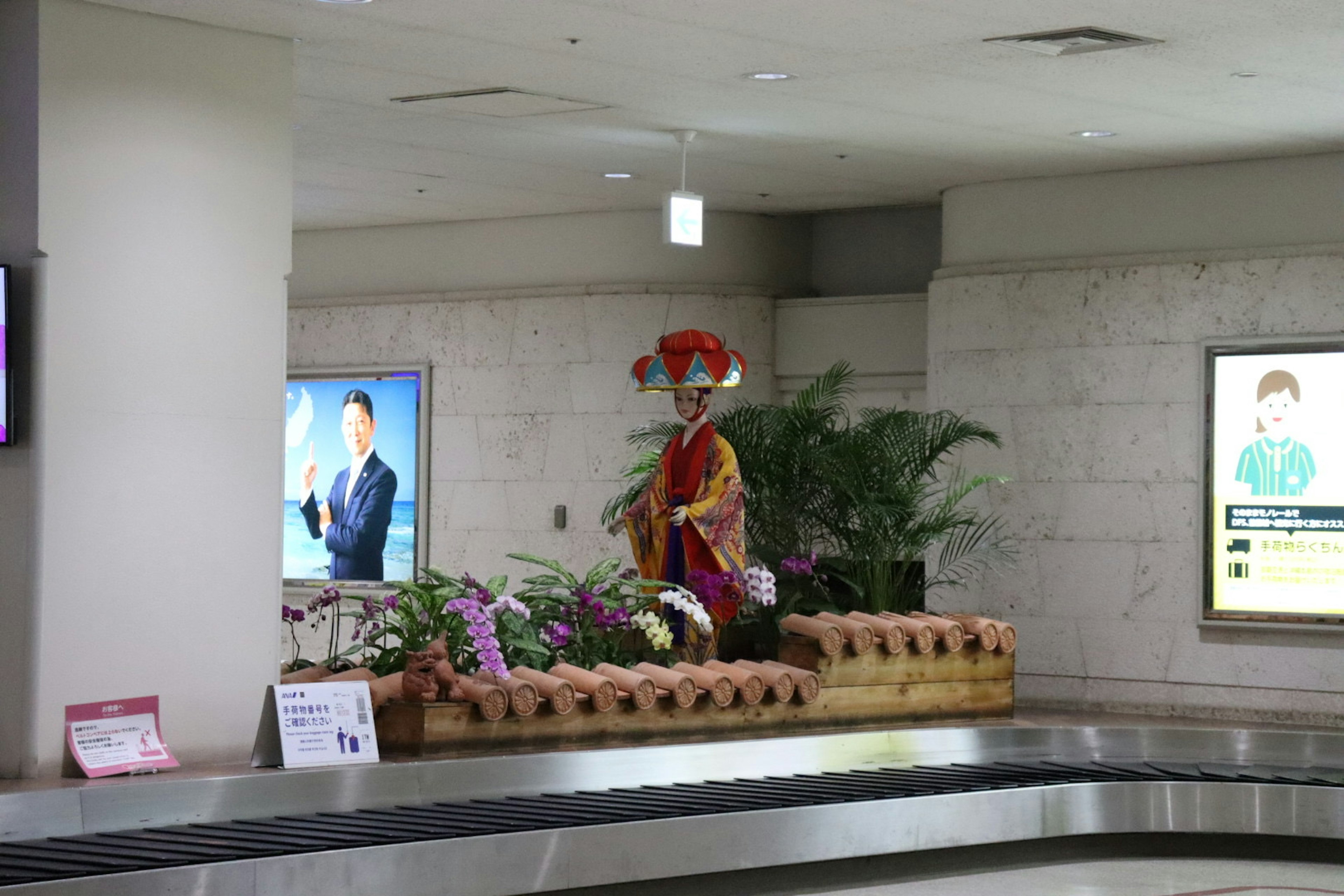 Une figure traditionnelle en costume entourée de plantes dans une zone de récupération des bagages d'aéroport