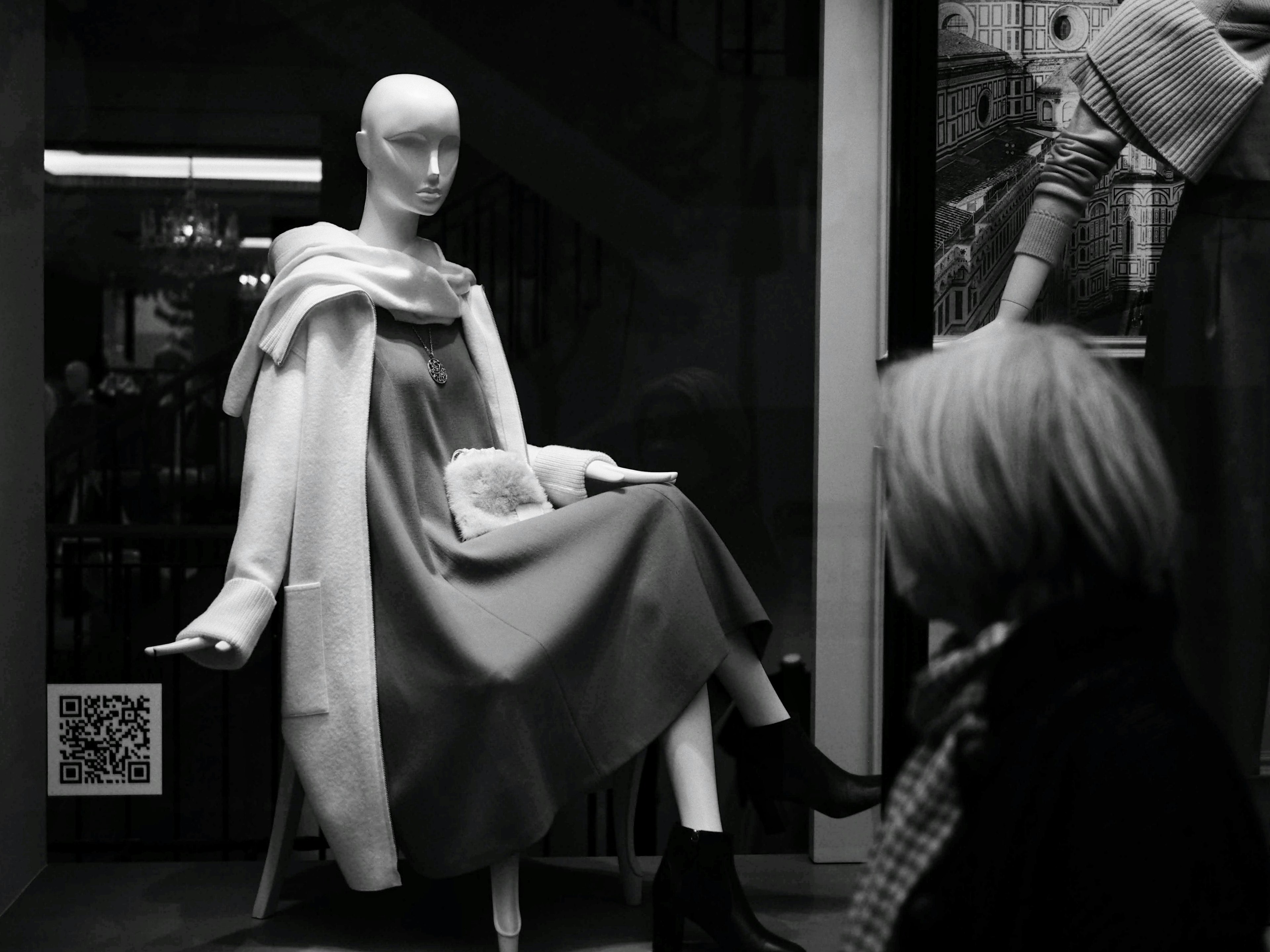 A mannequin seated in a black and white window display wearing a dress and coat