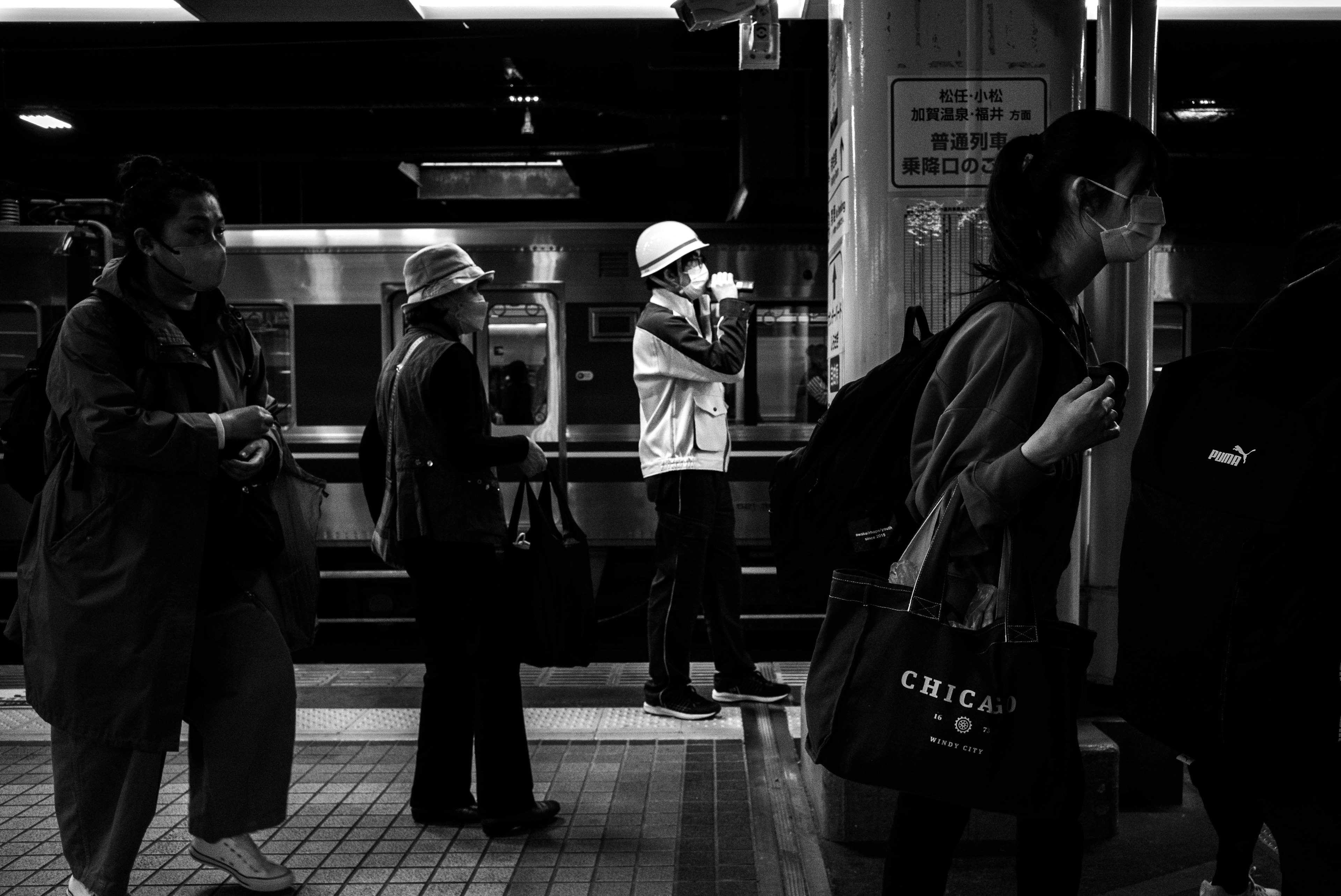 黒と白のモノクロ画像で、駅のホームに立つ人々が写っている