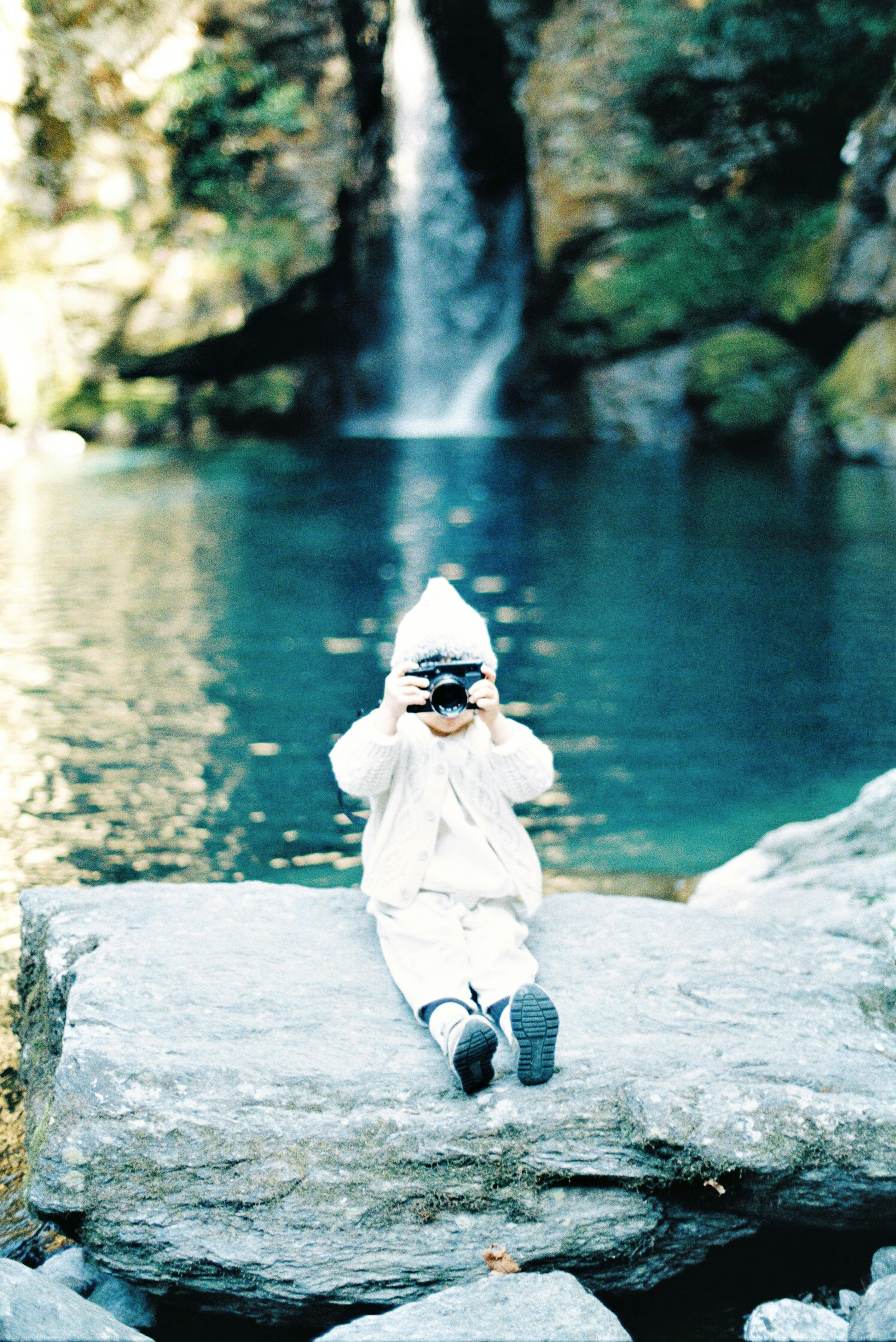 Enfant en vêtements blancs et chapeau prenant une photo devant une cascade avec de l'eau bleu clair