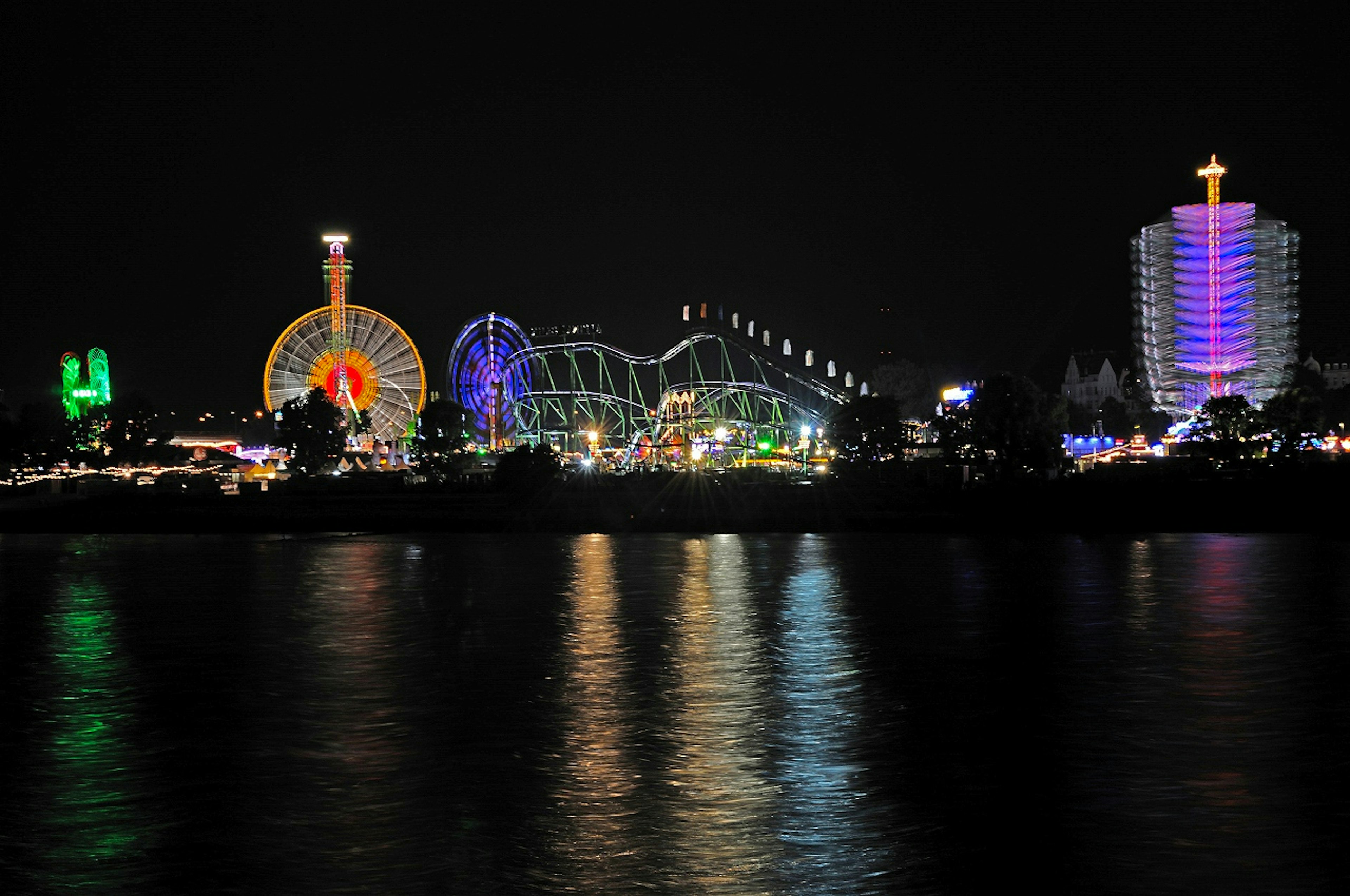 Nighttime view of a theme park with illuminated ferris wheel and roller coaster reflected on the water