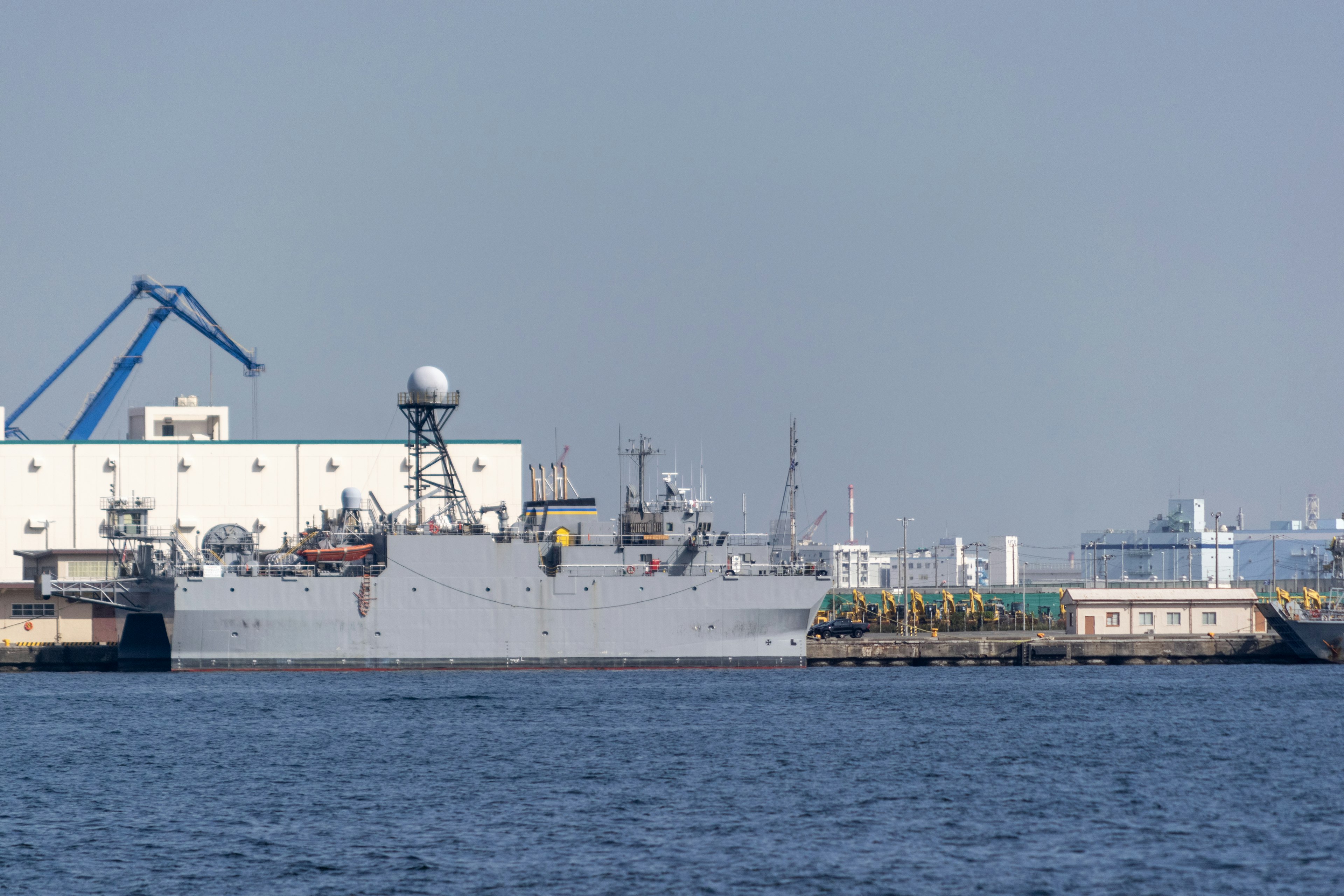 Military ship docked at the harbor with surrounding industrial buildings