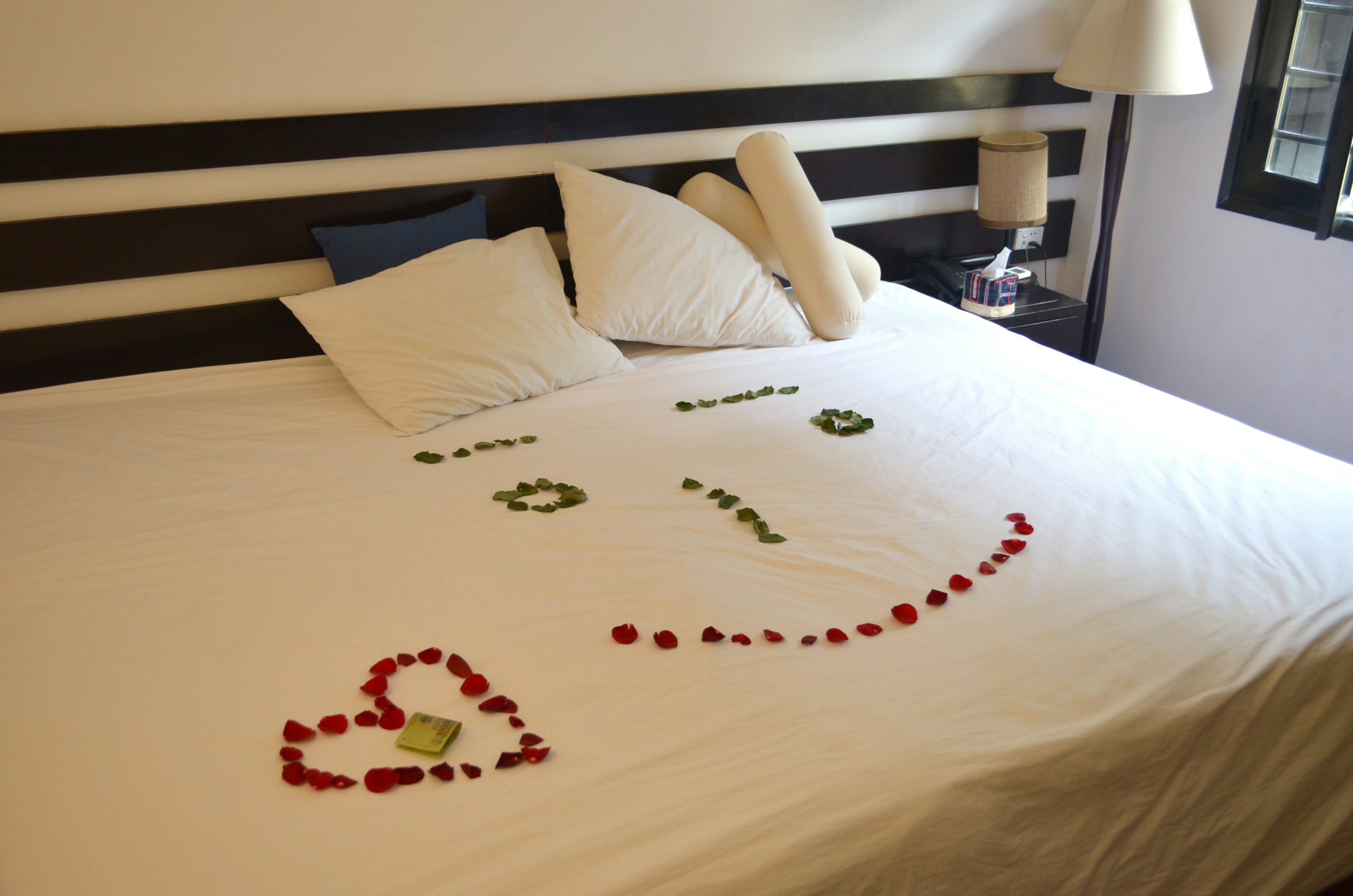 A bed with white sheets and pillows decorated with rose petals forming a smiley face and heart