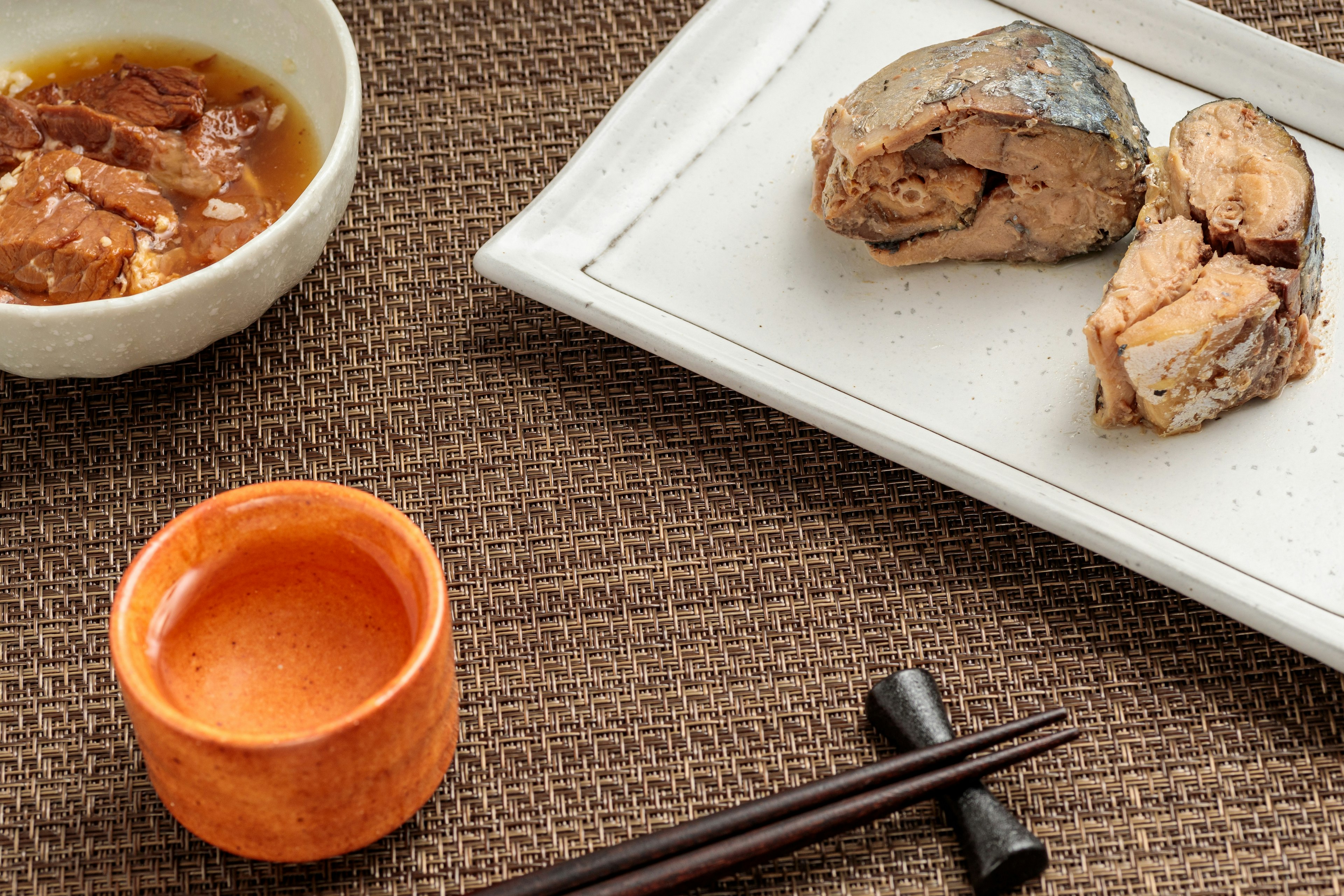 Un plato con filetes de pescado y una pequeña copa de sake sobre un mantel tejido