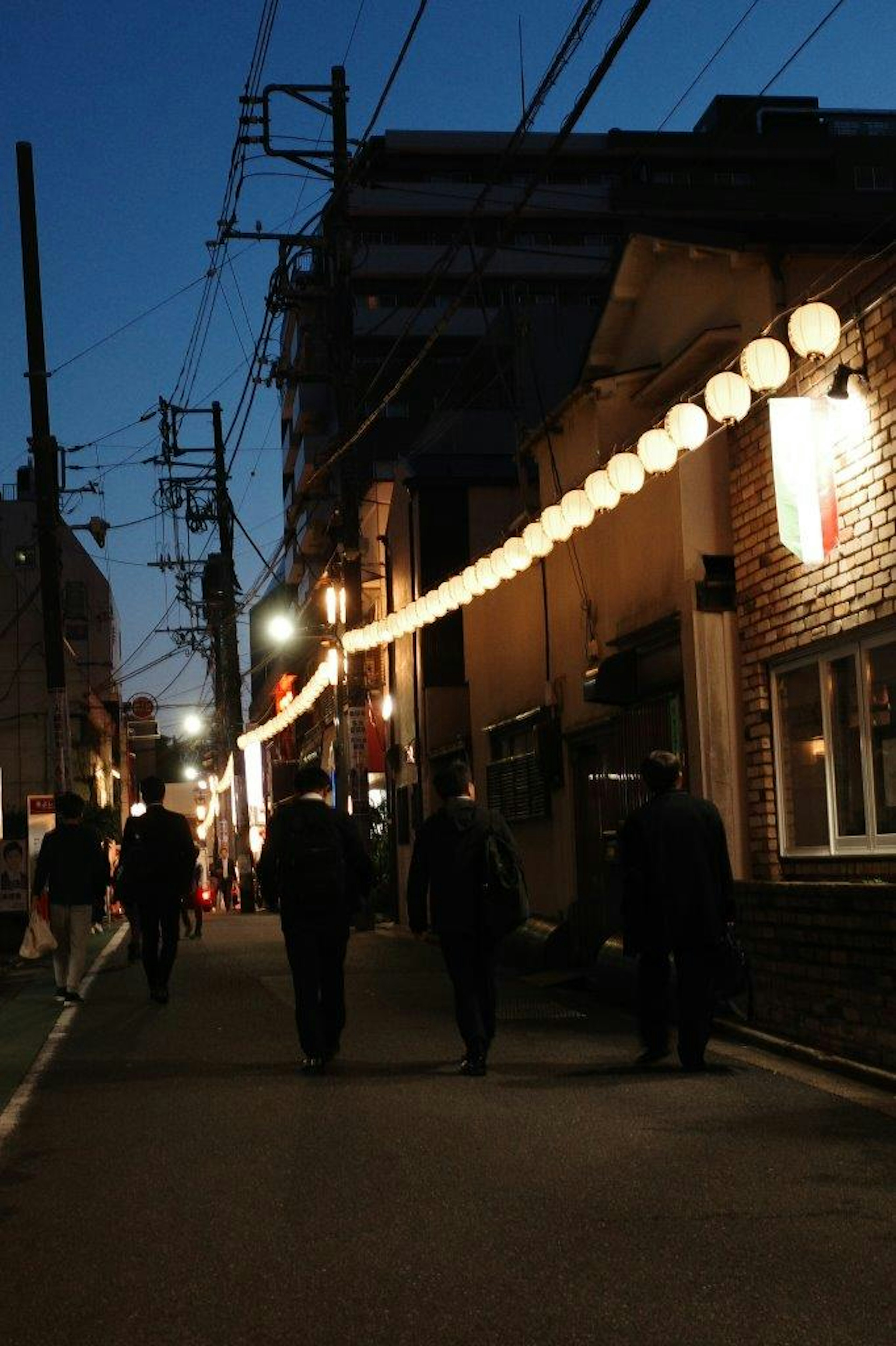 Escena de calle nocturna con personas caminando bajo luces de farol y edificios a lo largo de la calle