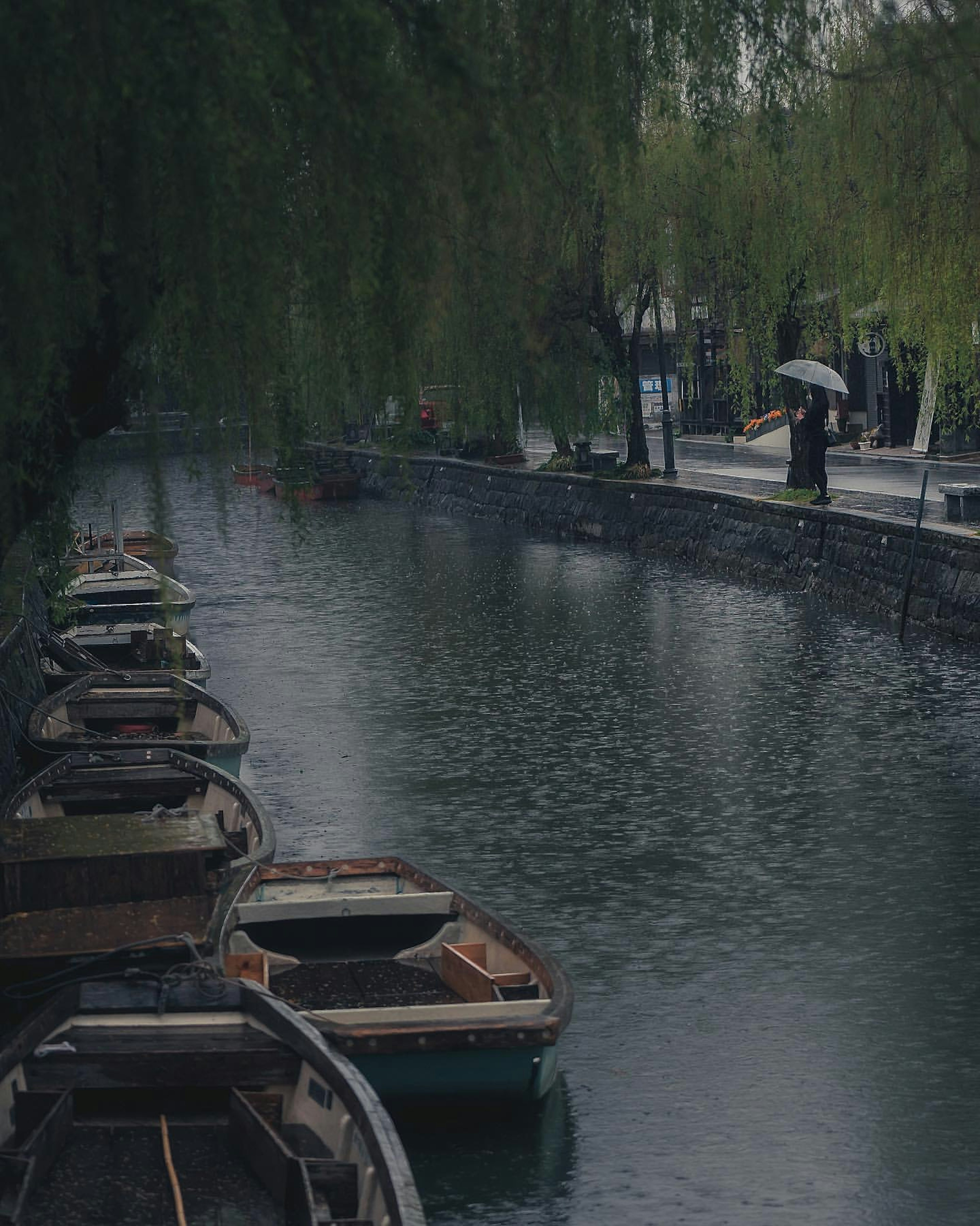 Canal tranquilo con pequeñas barcas alineadas bajo la lluvia y una persona caminando con un paraguas