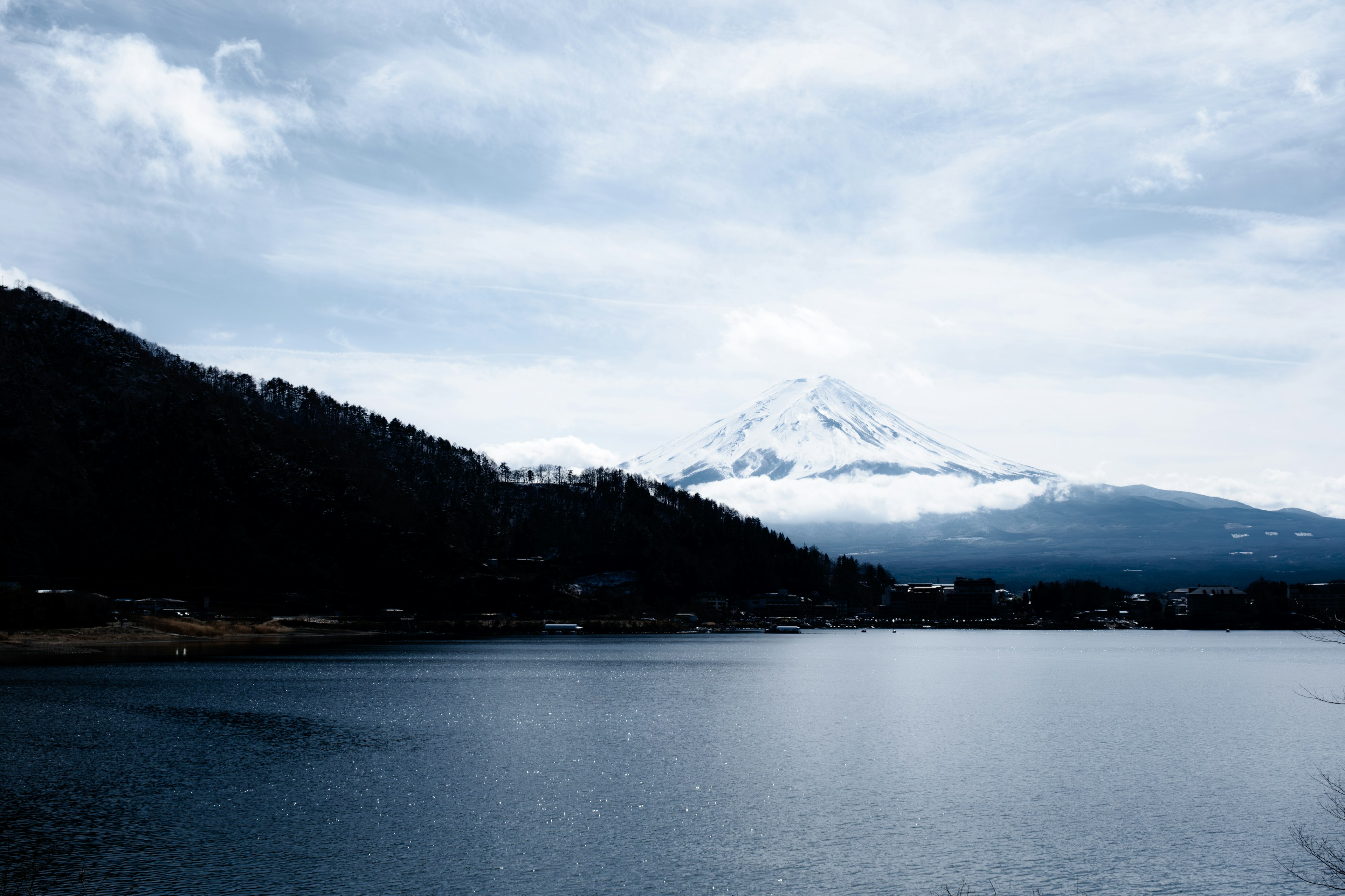 Snow-capped Mount Fuji shrouded in clouds with a serene lake in the foreground