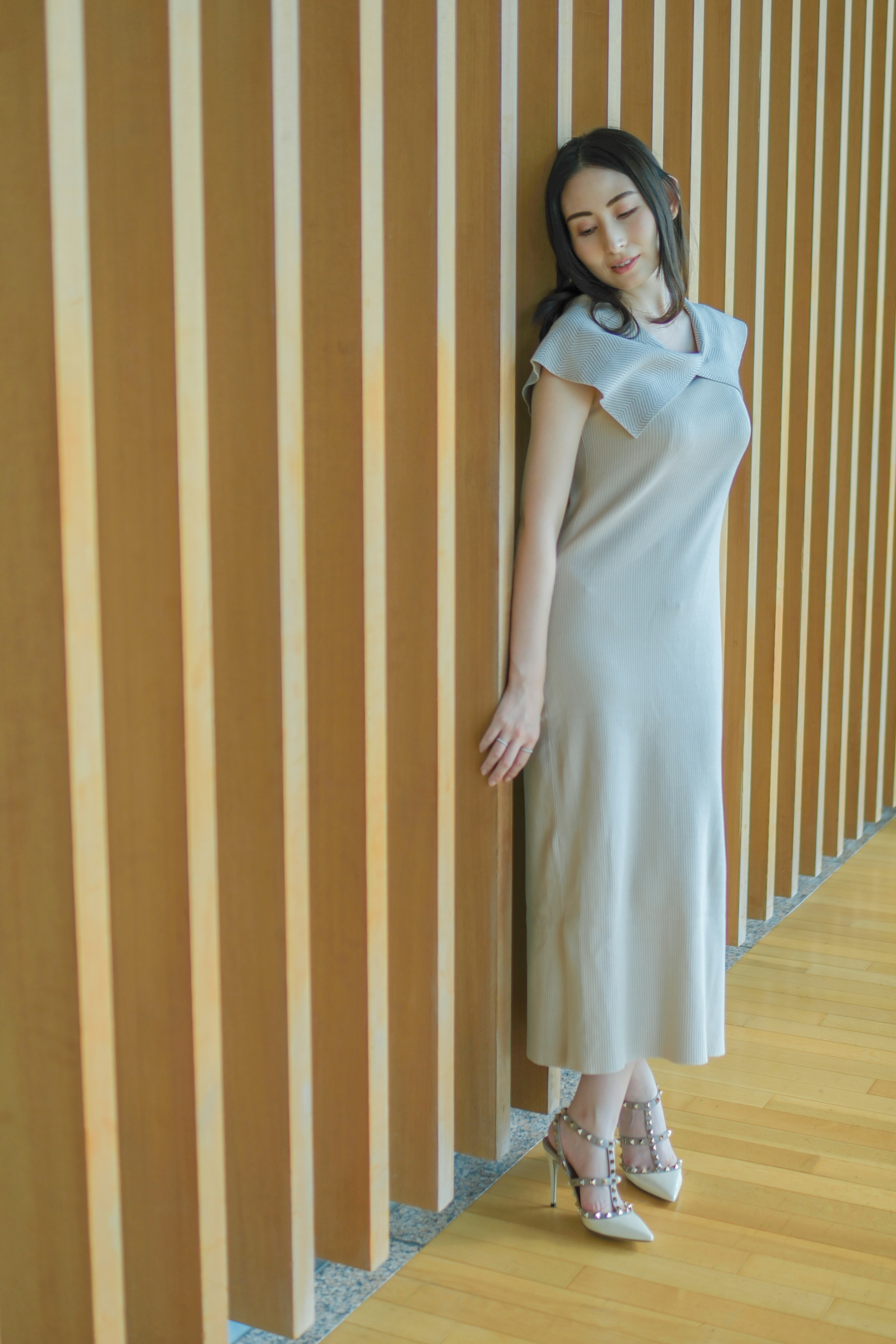 A woman leaning against a wooden wall wearing a light gray dress