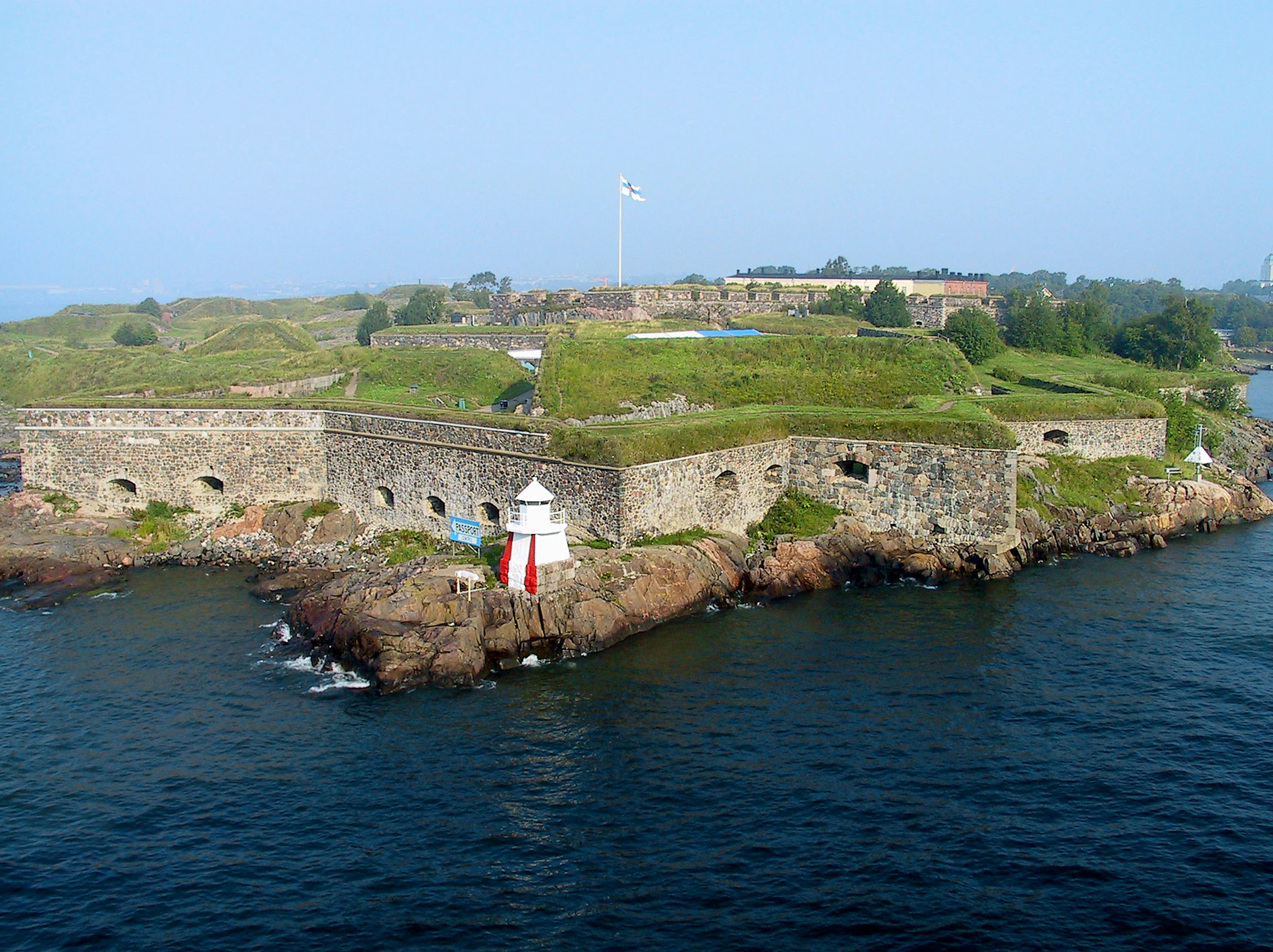 Historic coastal fortress surrounded by water featuring green hills and a white lighthouse