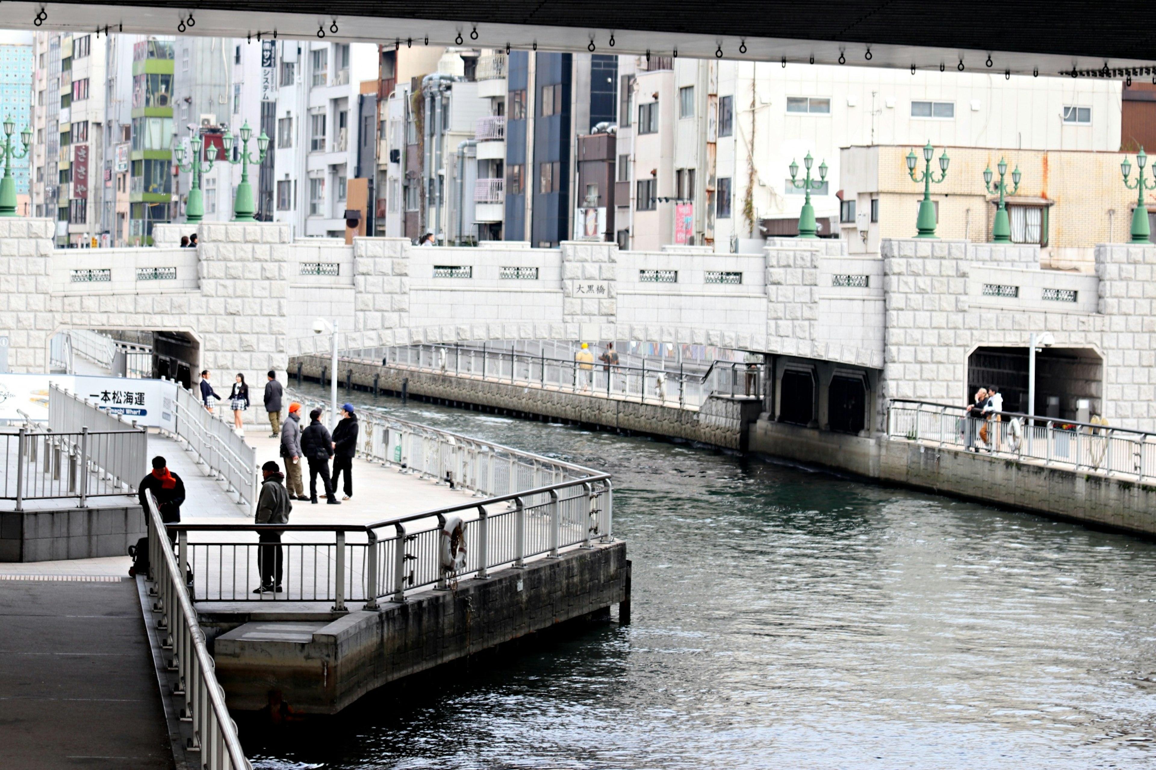 Personas reunidas en un paseo junto al río con edificios al fondo