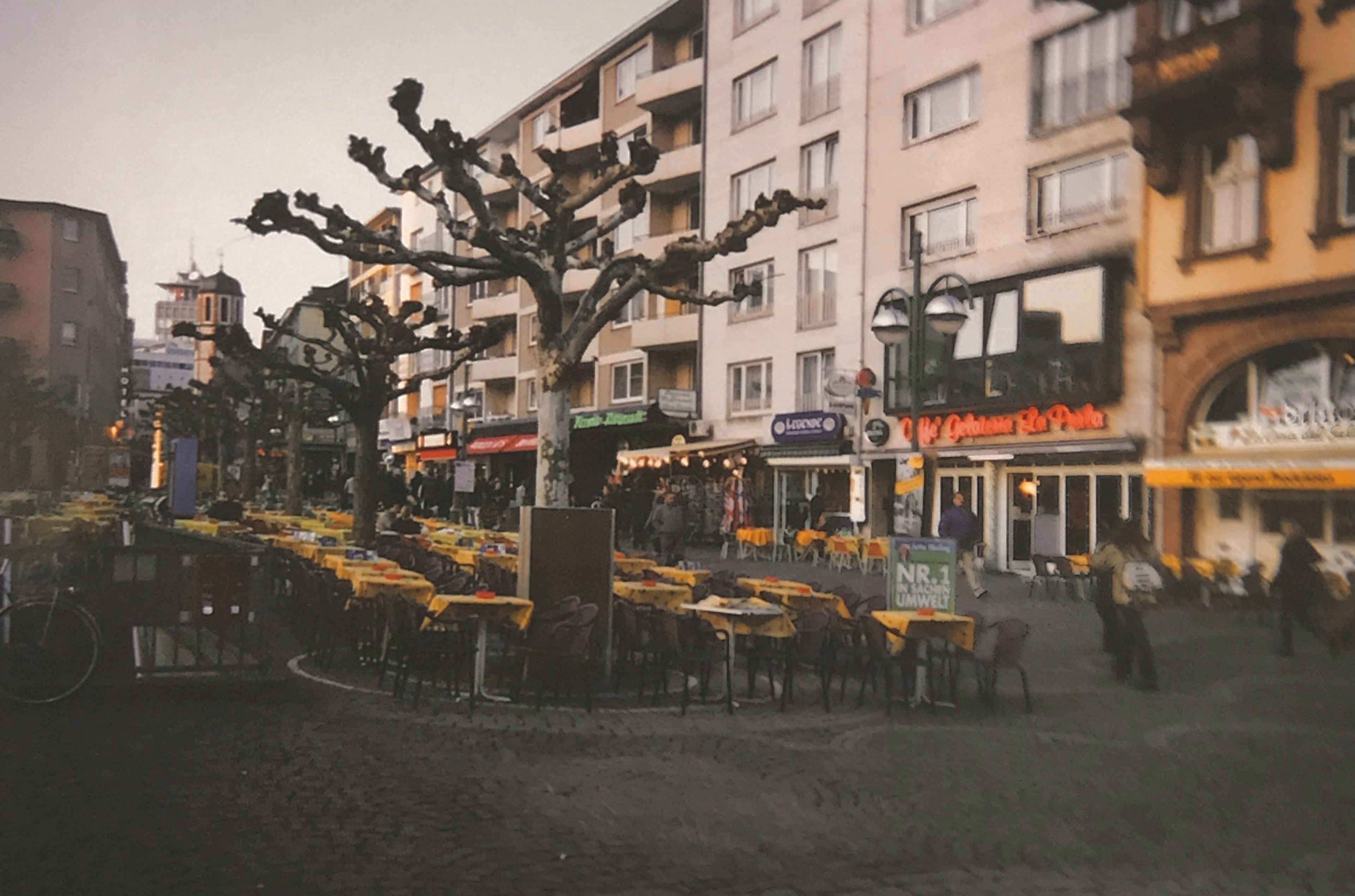 Una escena de esquina de calle con cafés y restaurantes, mesas y sillas dispuestas en una plaza con árboles podados