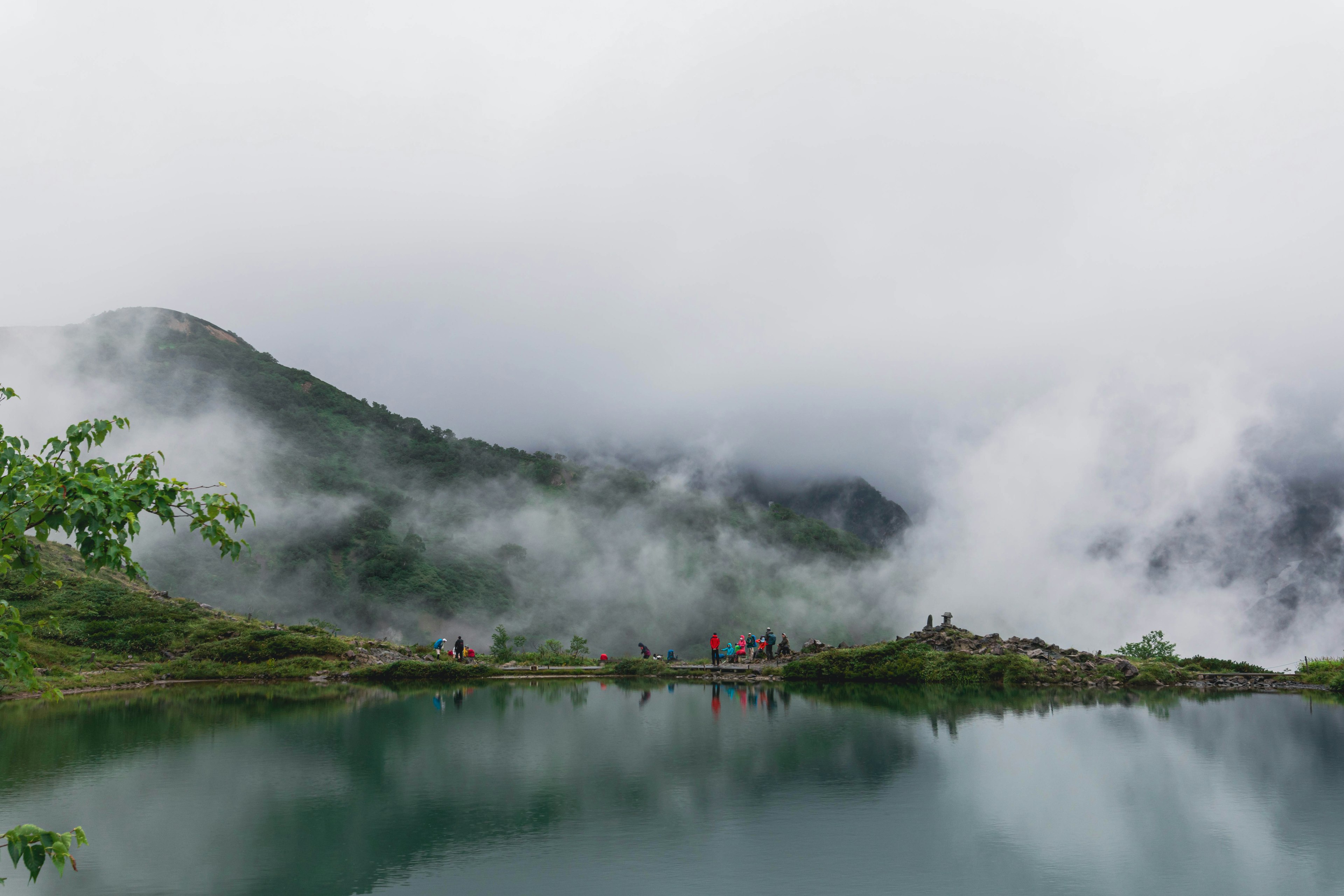 Paesaggio di montagne nebbiose e lago calmo con persone che attraversano un ponte