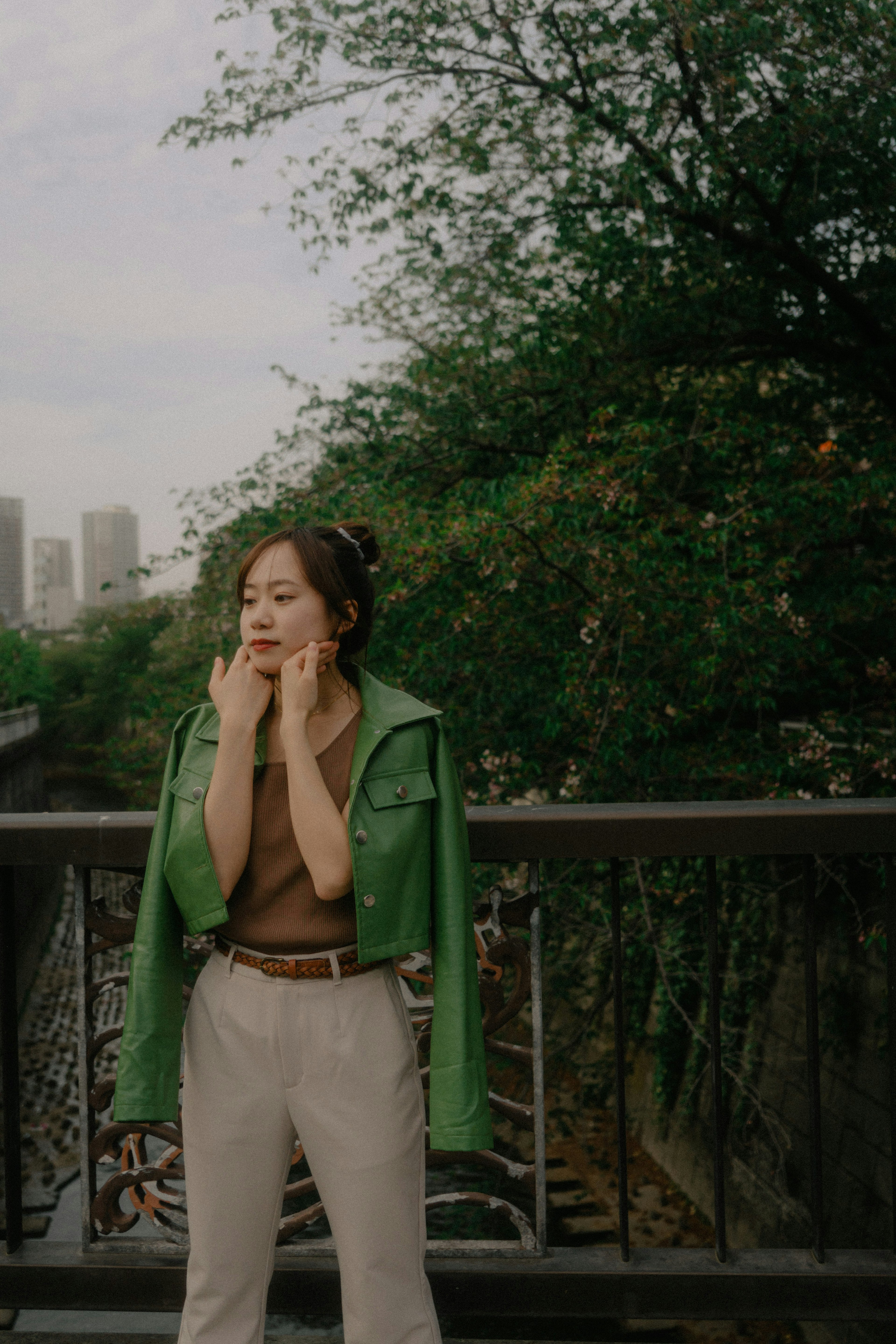 A woman in a green jacket stands on a bridge with greenery in the background