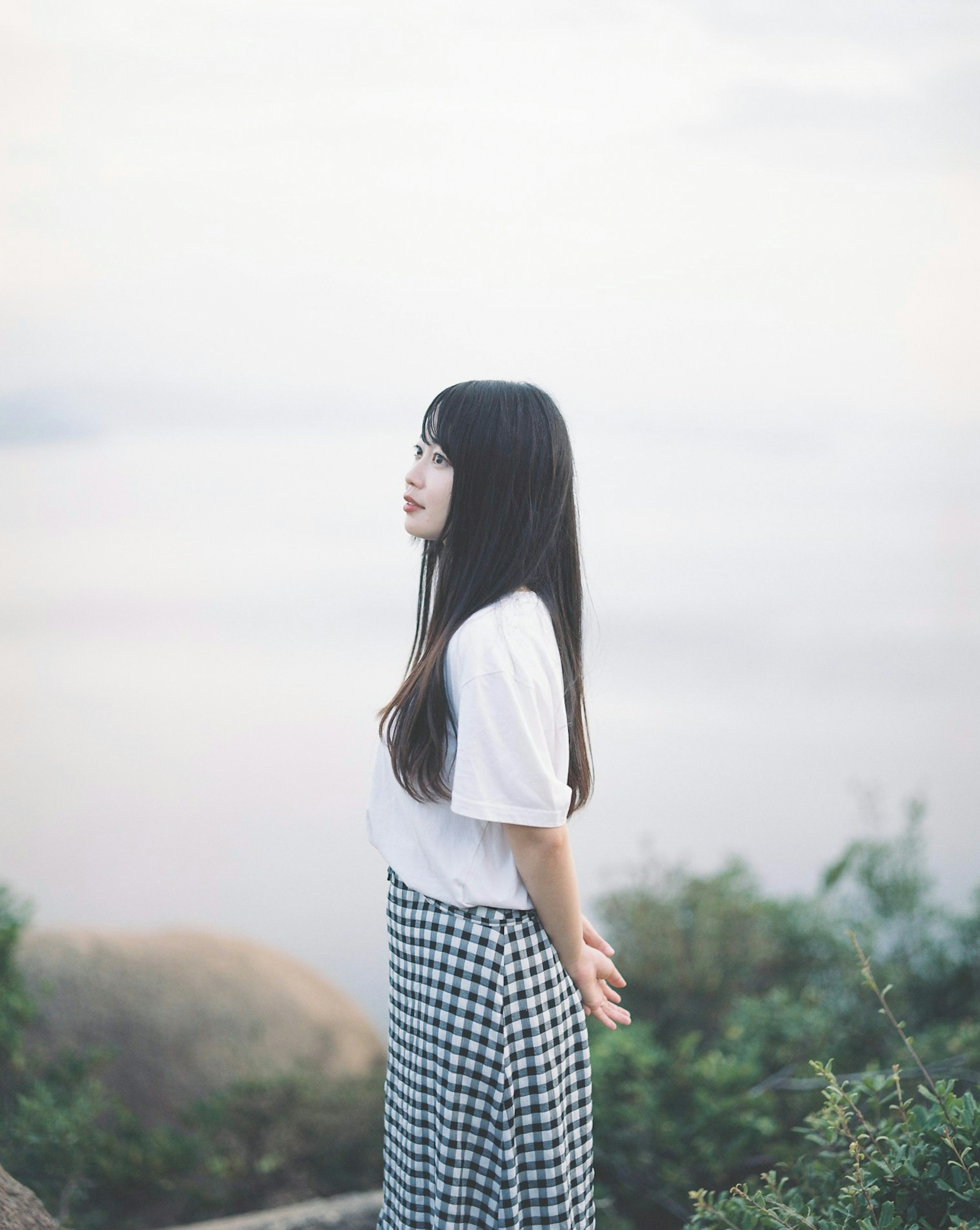 A woman standing quietly by a lake wearing a white t-shirt and a checkered skirt