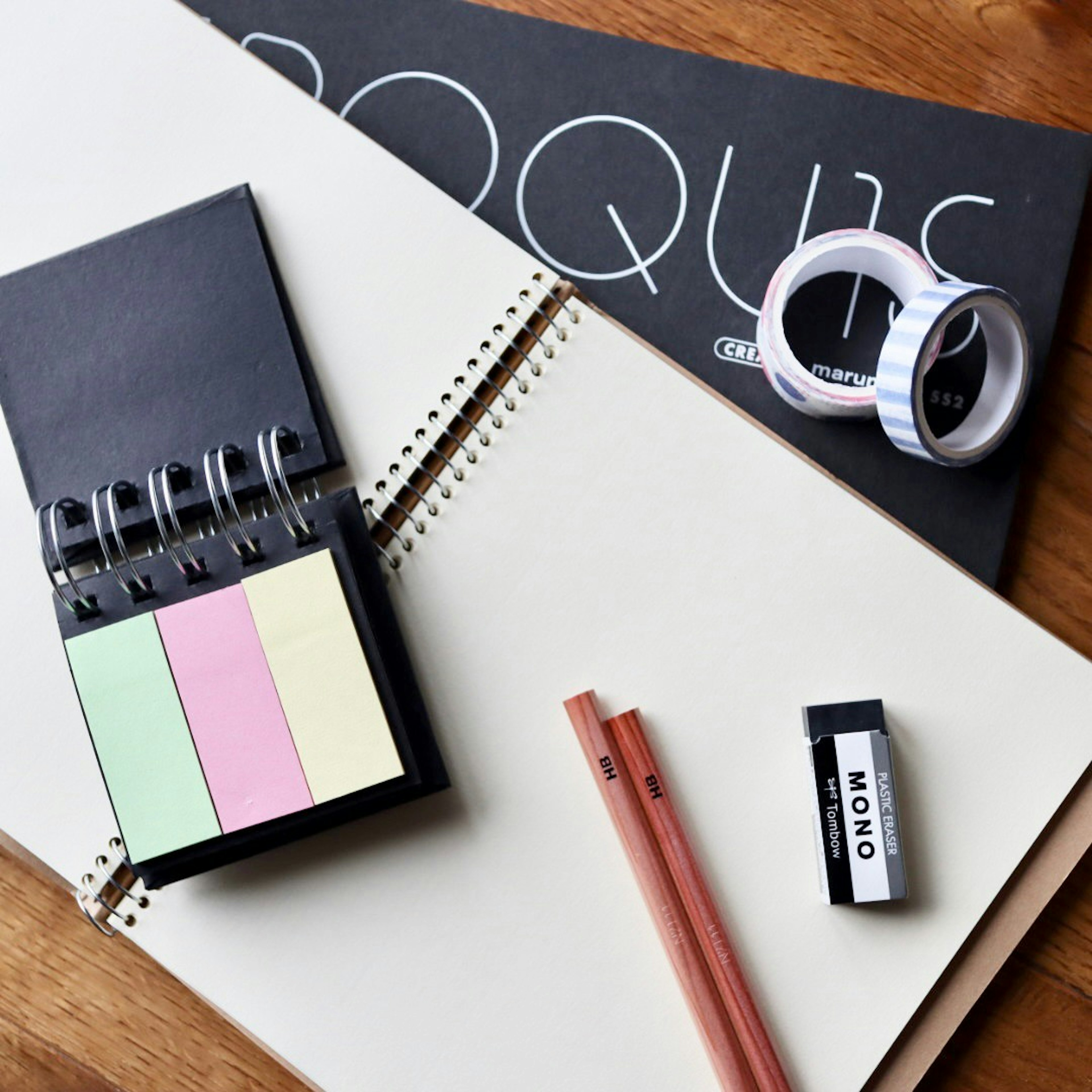 An arrangement of stationery items including a spiral notebook colorful sticky notes red pencils and white tape