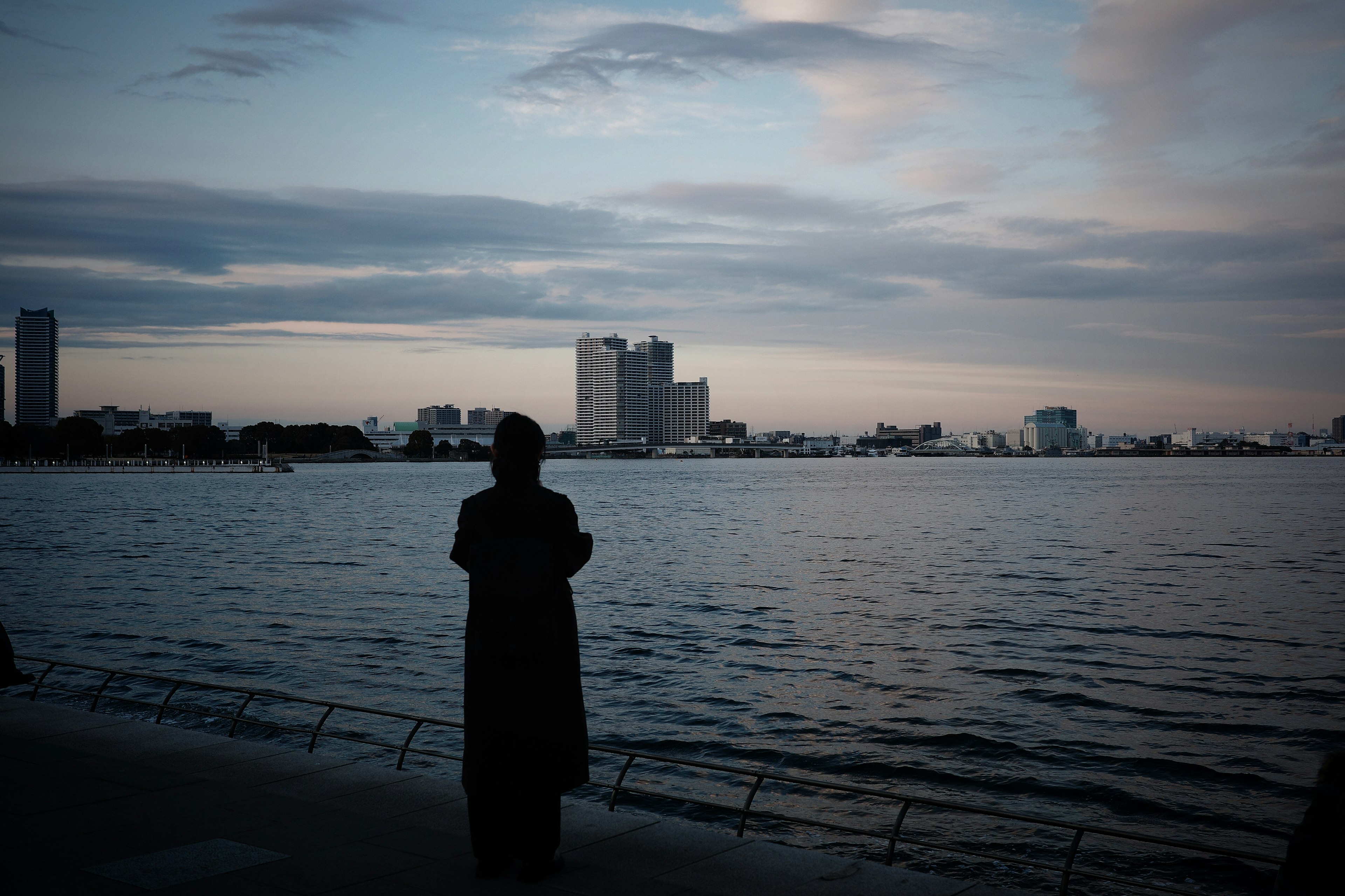 Eine Figur steht am Wasser und beobachtet den Sonnenuntergang über der Stadt