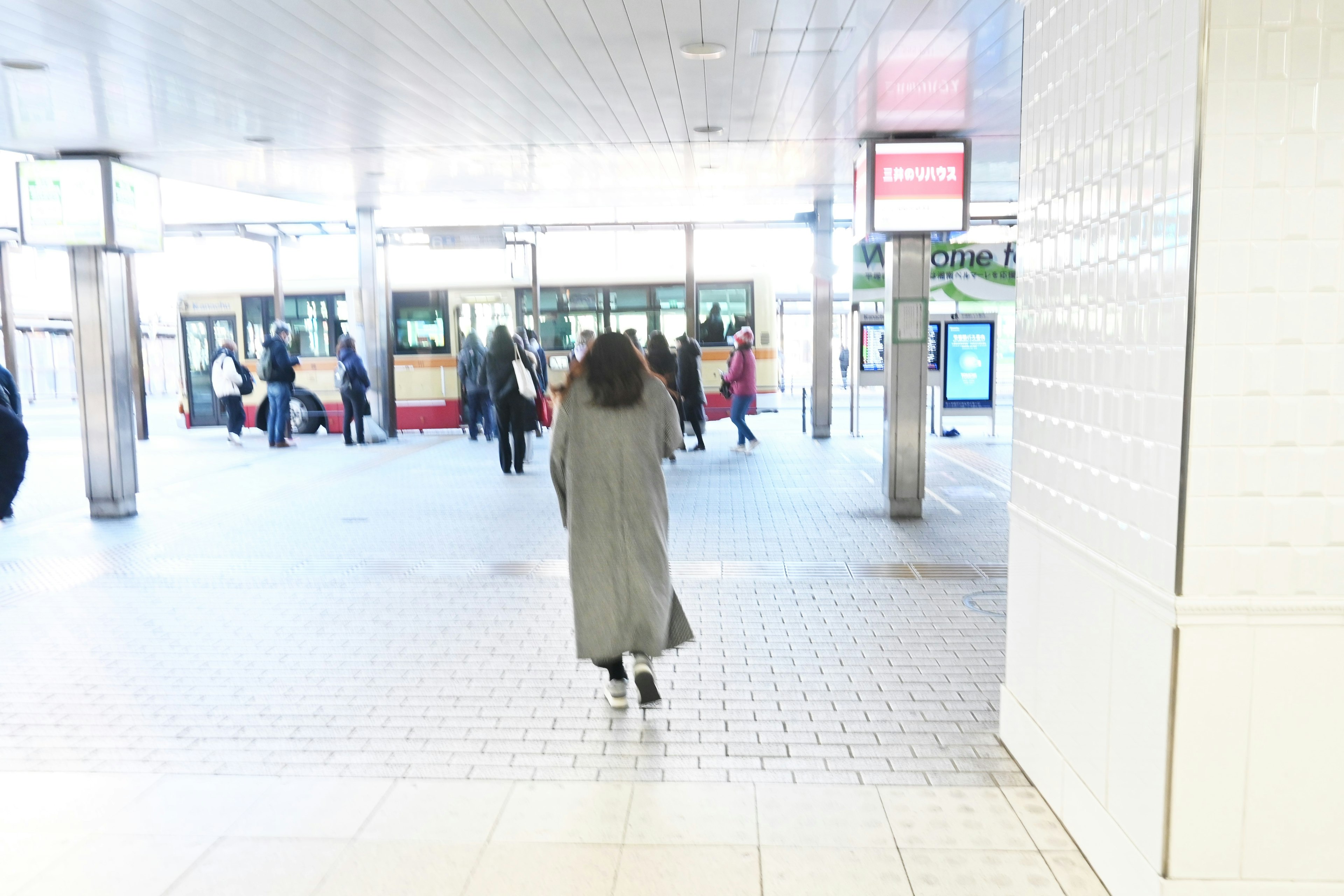 A bright train station interior with people walking and a woman in a gray coat