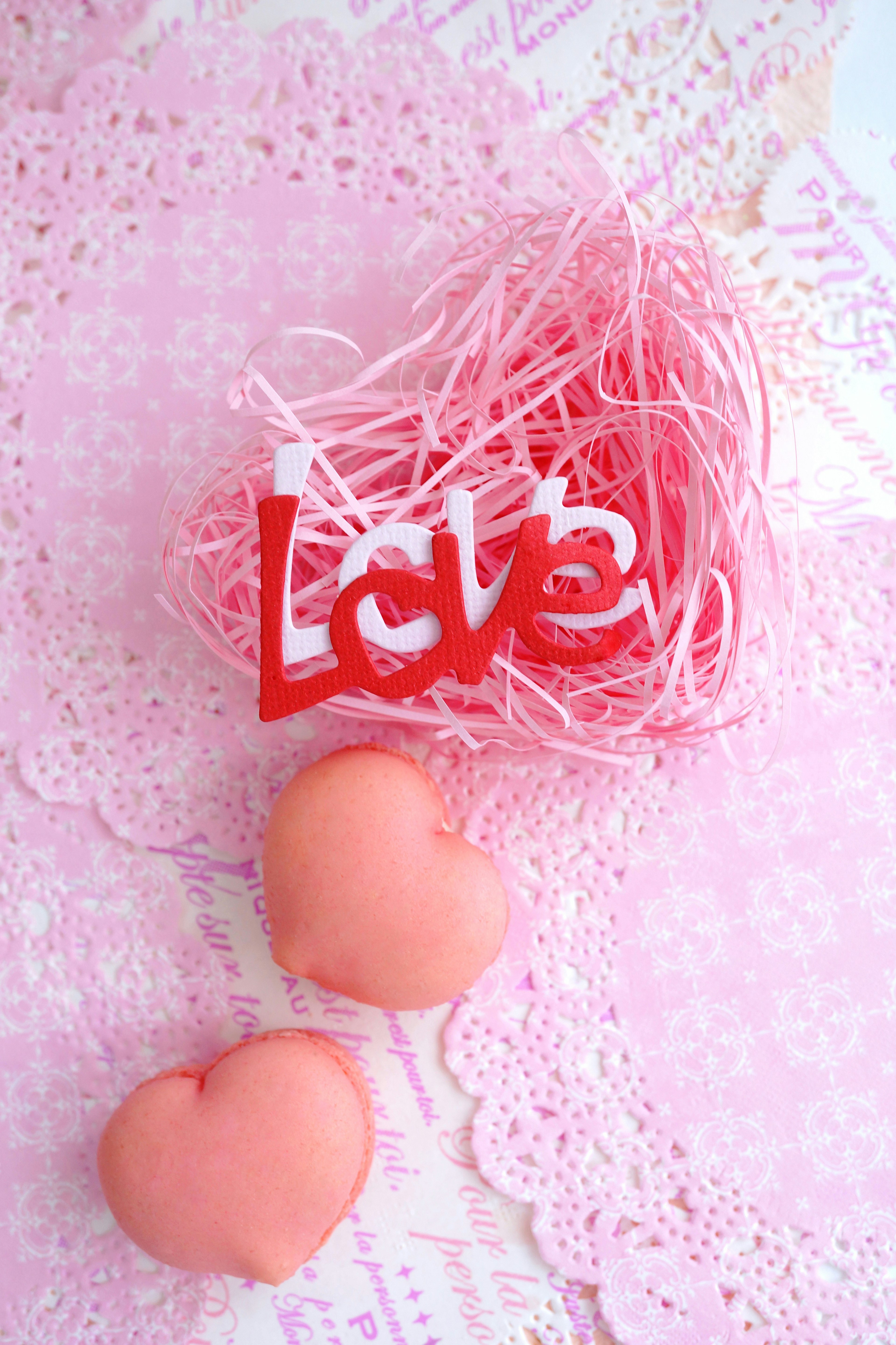 Red heart-shaped 'Love' text on a pink background with soft heart-shaped candies and decorative lace