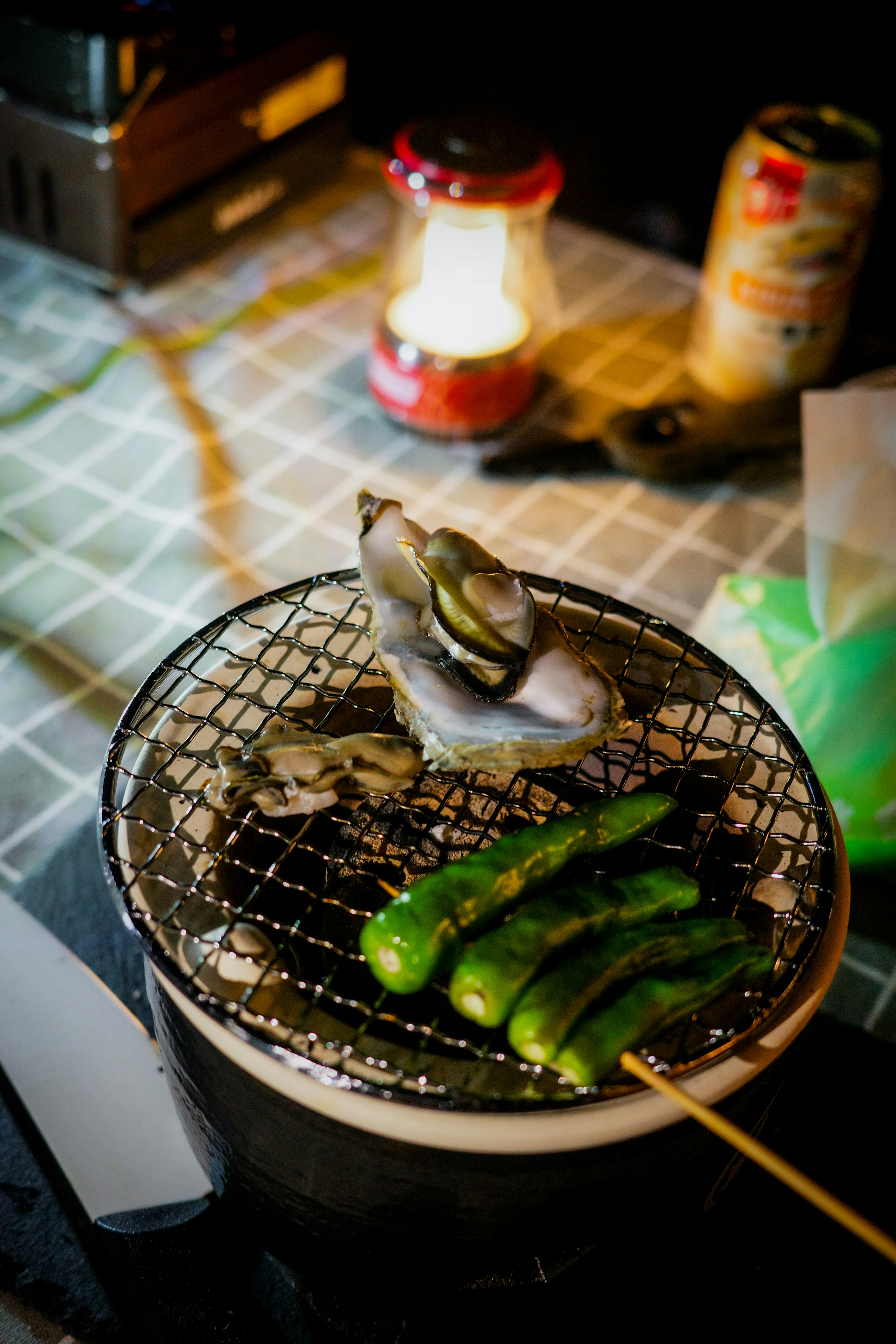 그릴에 조개와 청양고추 꼬치가 놓여 있고 저녁 불빛 아래 랜턴과 맥주 캔이 배경에 있음