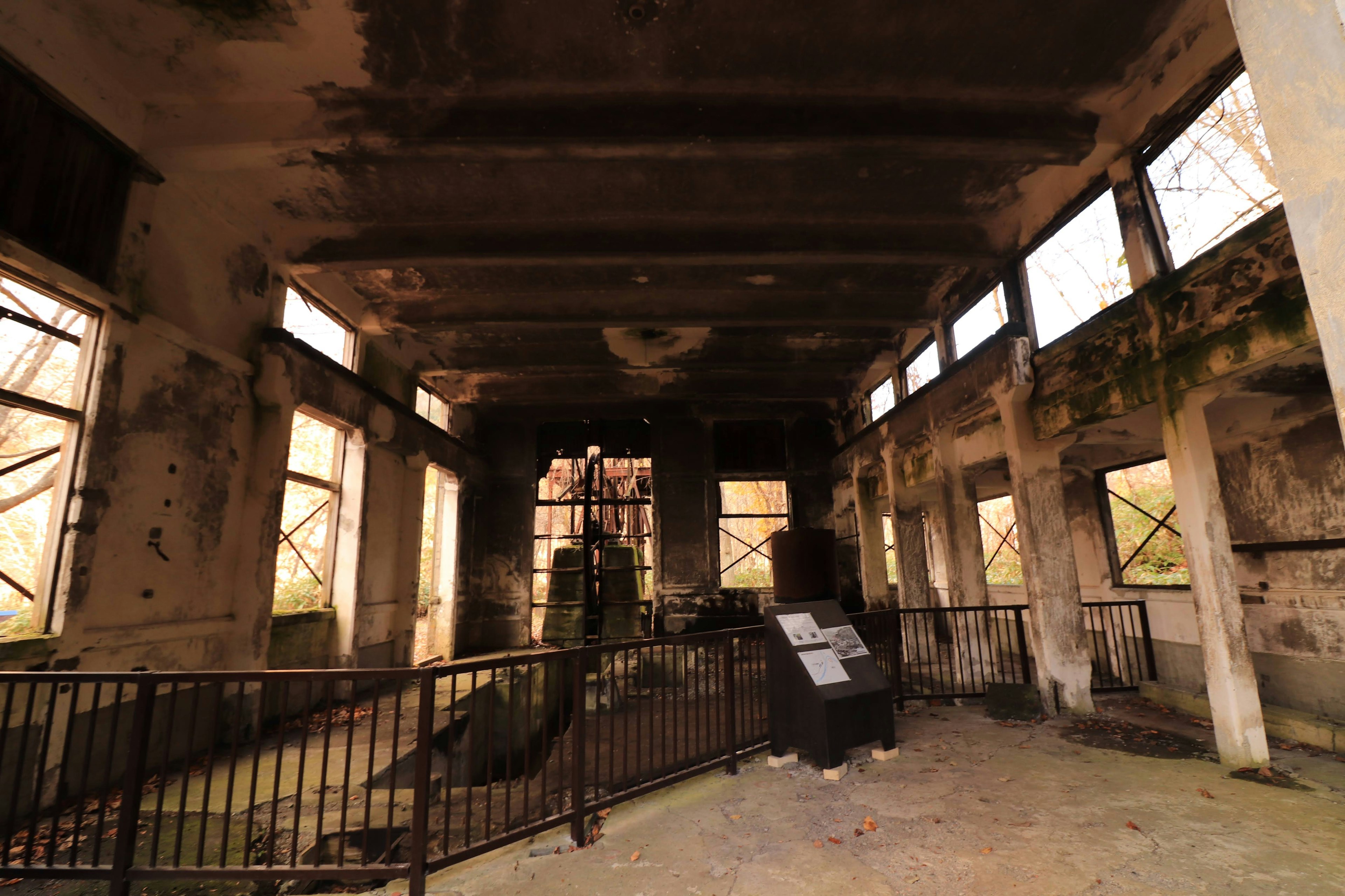 Interior de una fábrica en ruinas con paredes en mal estado y ventanas abiertas, barandilla de metal visible