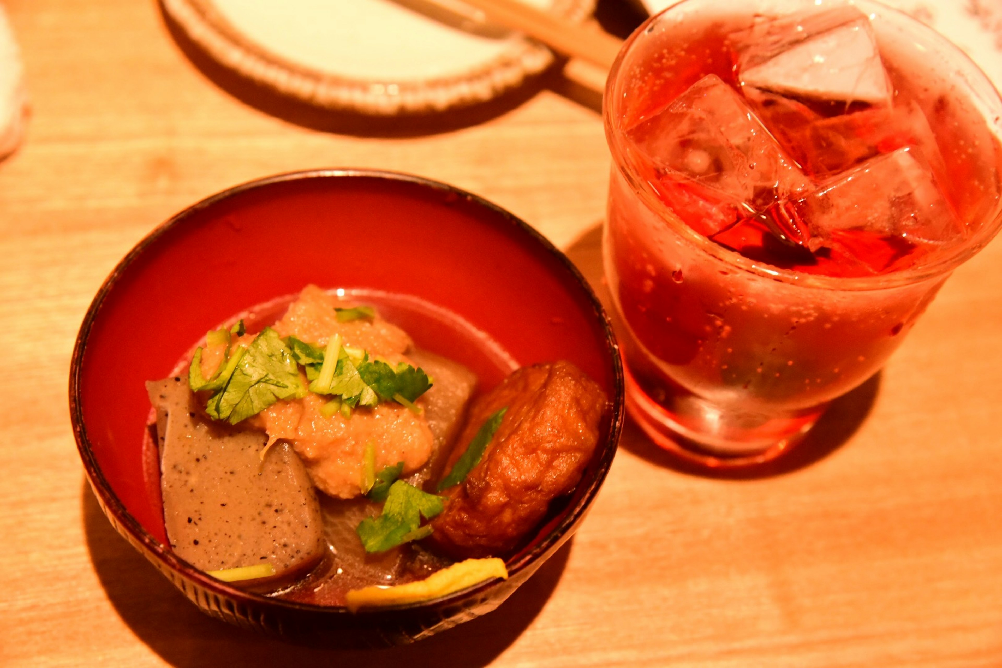 A red bowl of simmered dish with green garnish next to a glass of red drink with ice