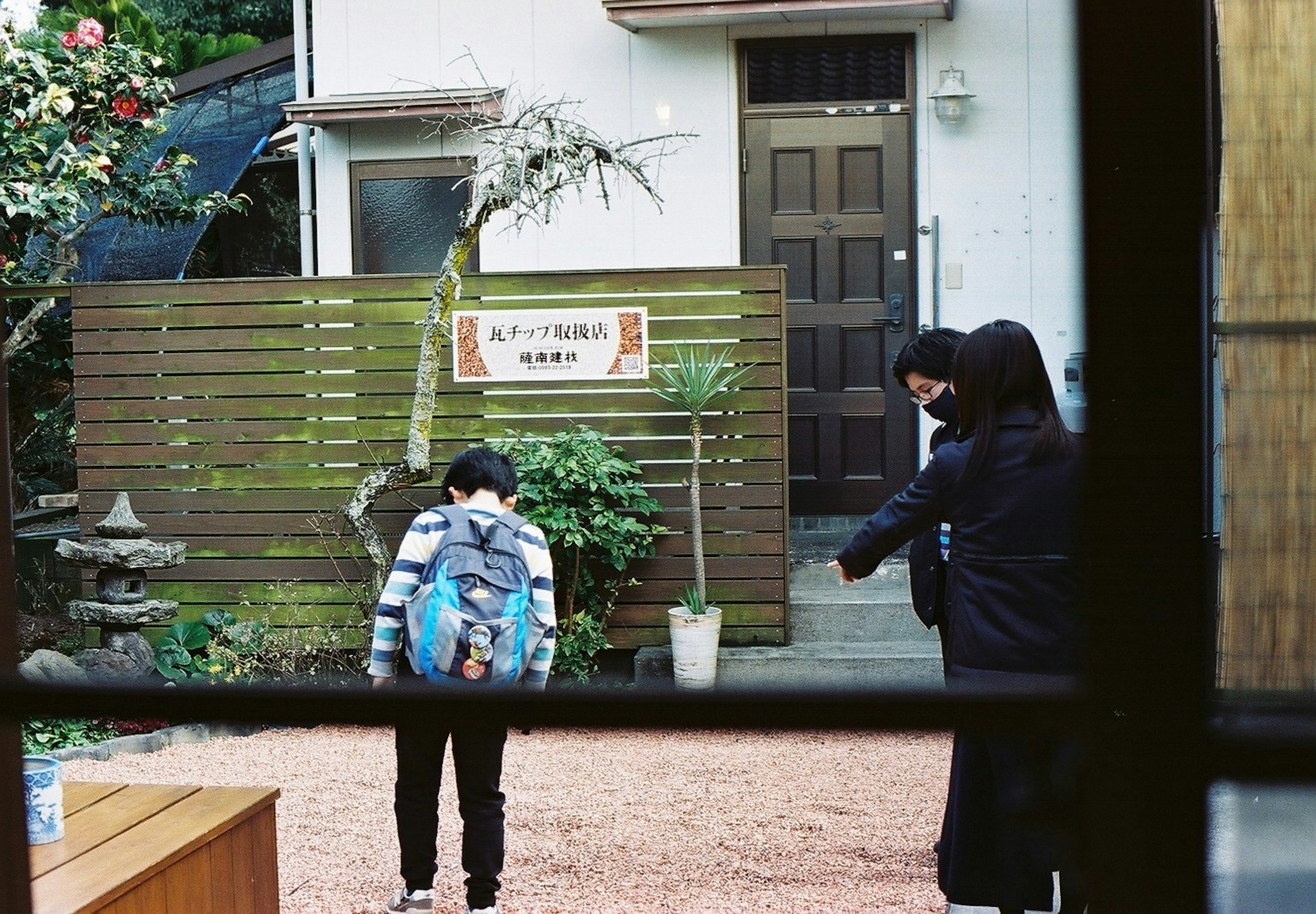 Ein Kind und ein Erwachsener sprechen in einem Garten, sichtbar durch ein Fenster, das ein japanisches Haus und Pflanzen zeigt