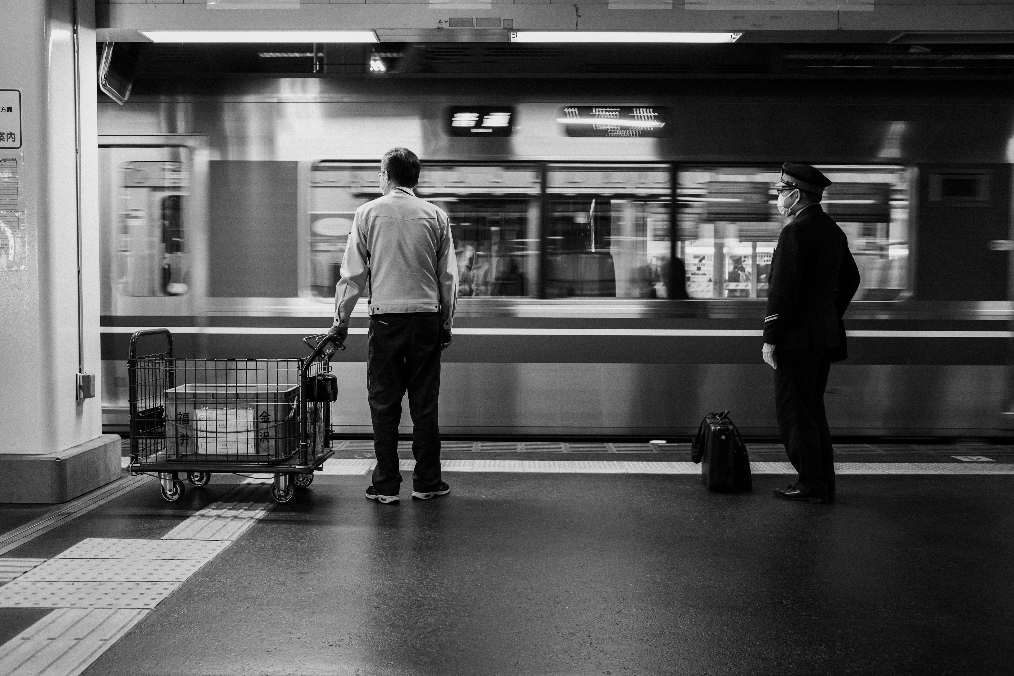 Two men waiting at a black and white train station with a cart