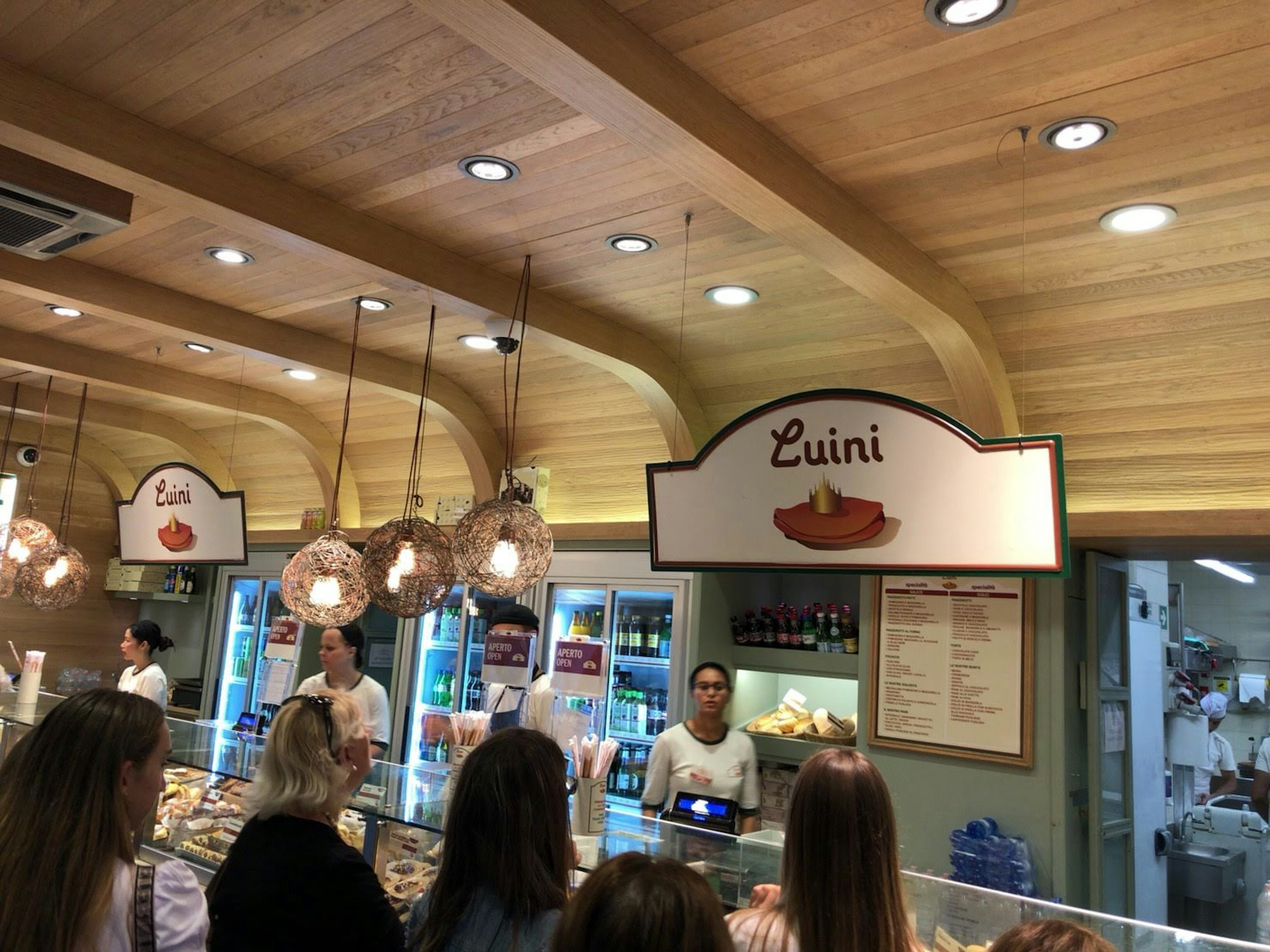 Clientes interactuando con el personal de la panadería Luini con interior de madera y luces colgantes