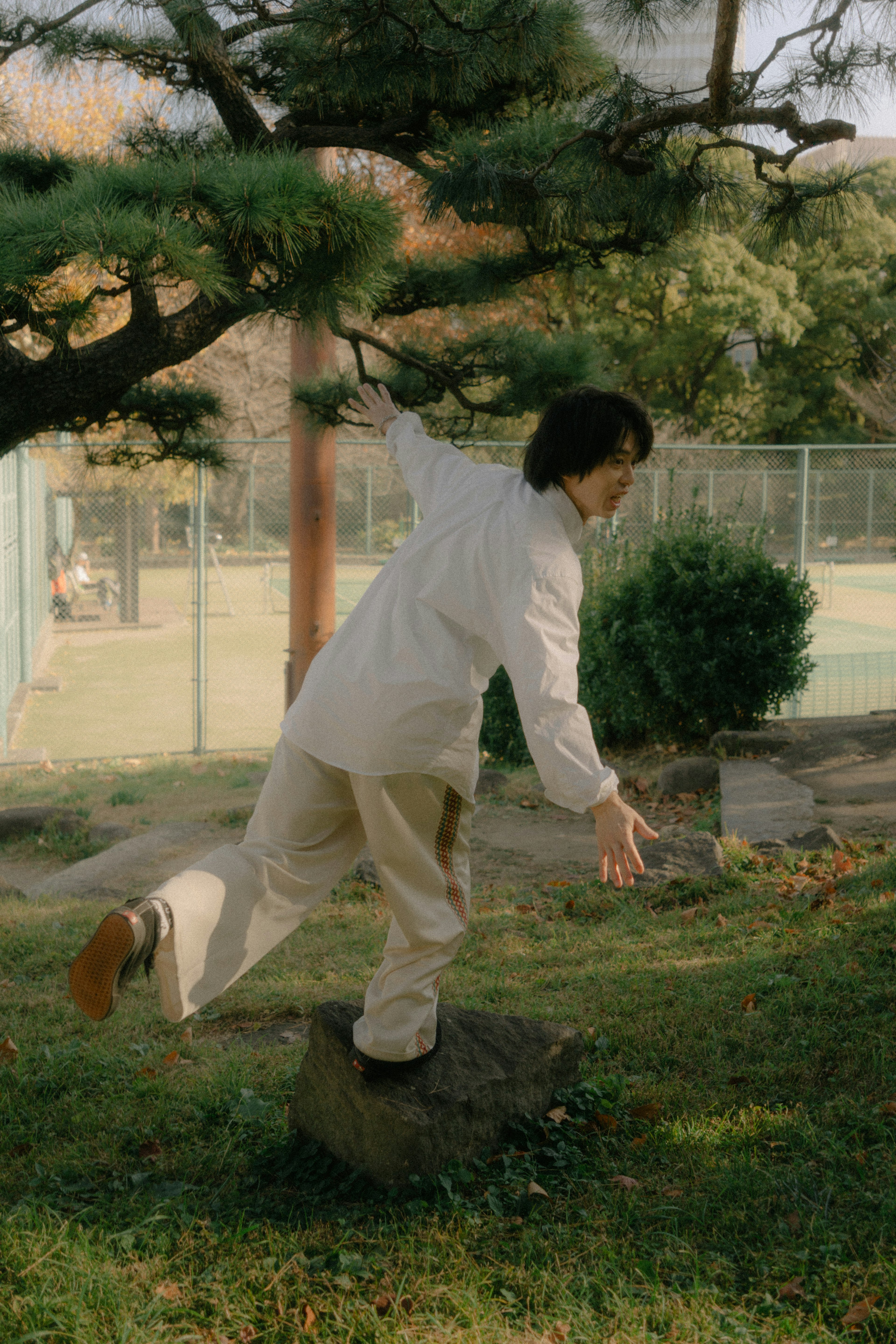 Person balancing on a stone in a park wearing a white shirt and beige pants surrounded by greenery