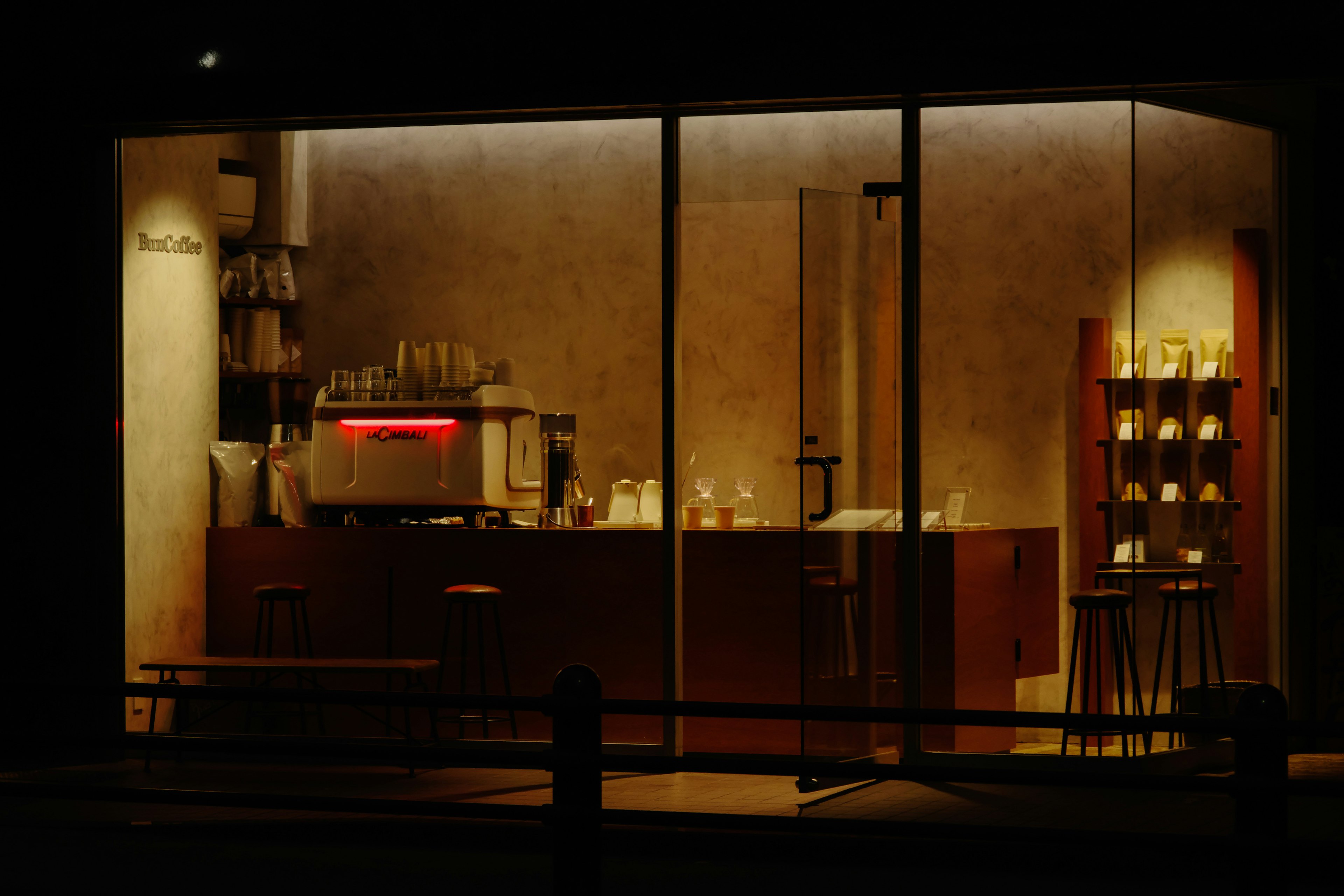 Vista interior de una cafetería a través de una ventana en un entorno oscuro con decoración moderna y una máquina de café