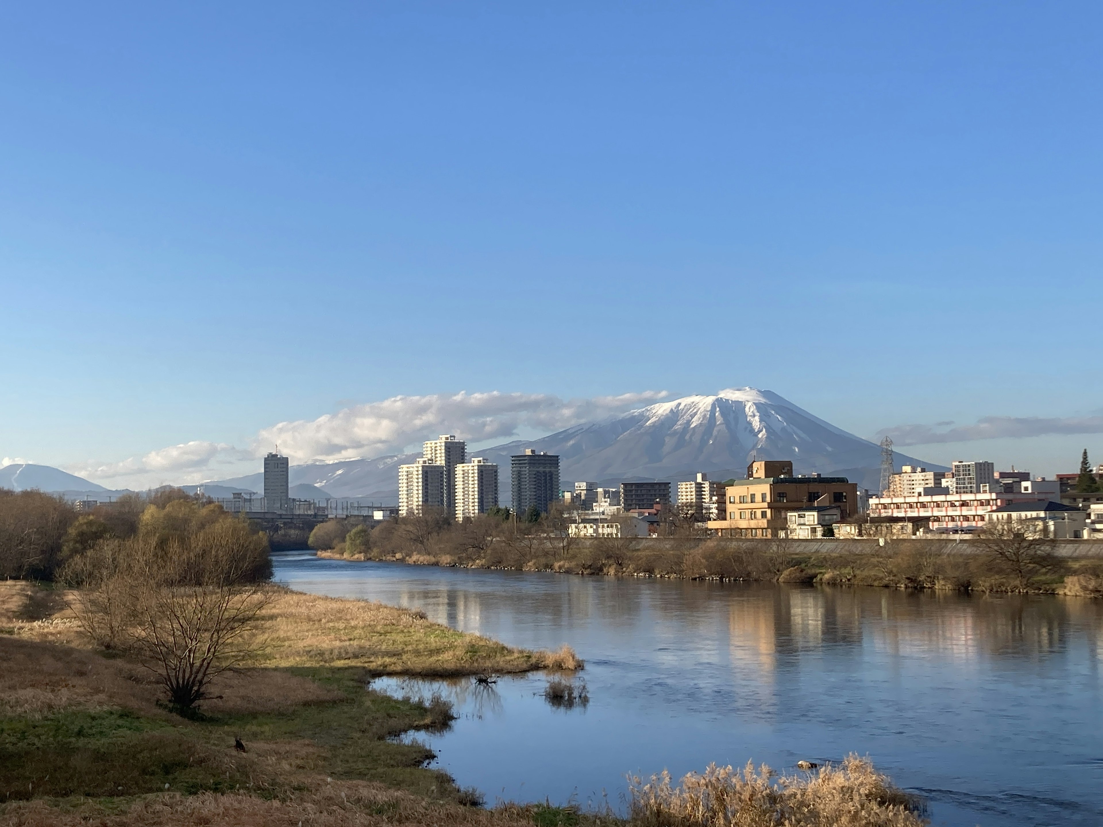 青空の下に広がる川と山の景色 近くには都市の建物が並び 雪をかぶった山が背景に見える