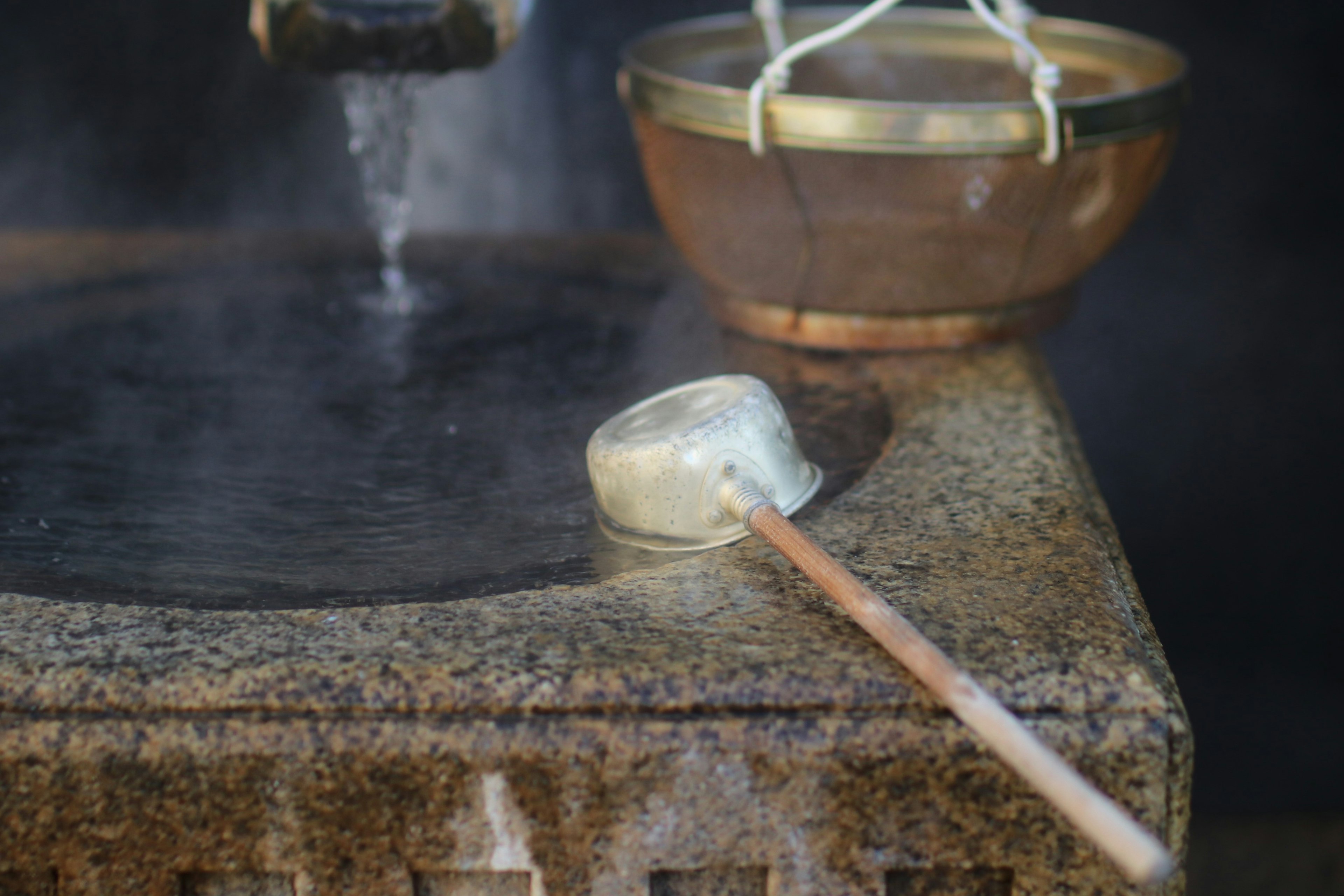 Image of a white bowl and wooden ladle placed on a warm stone with steam rising in the background and a metal bowl visible