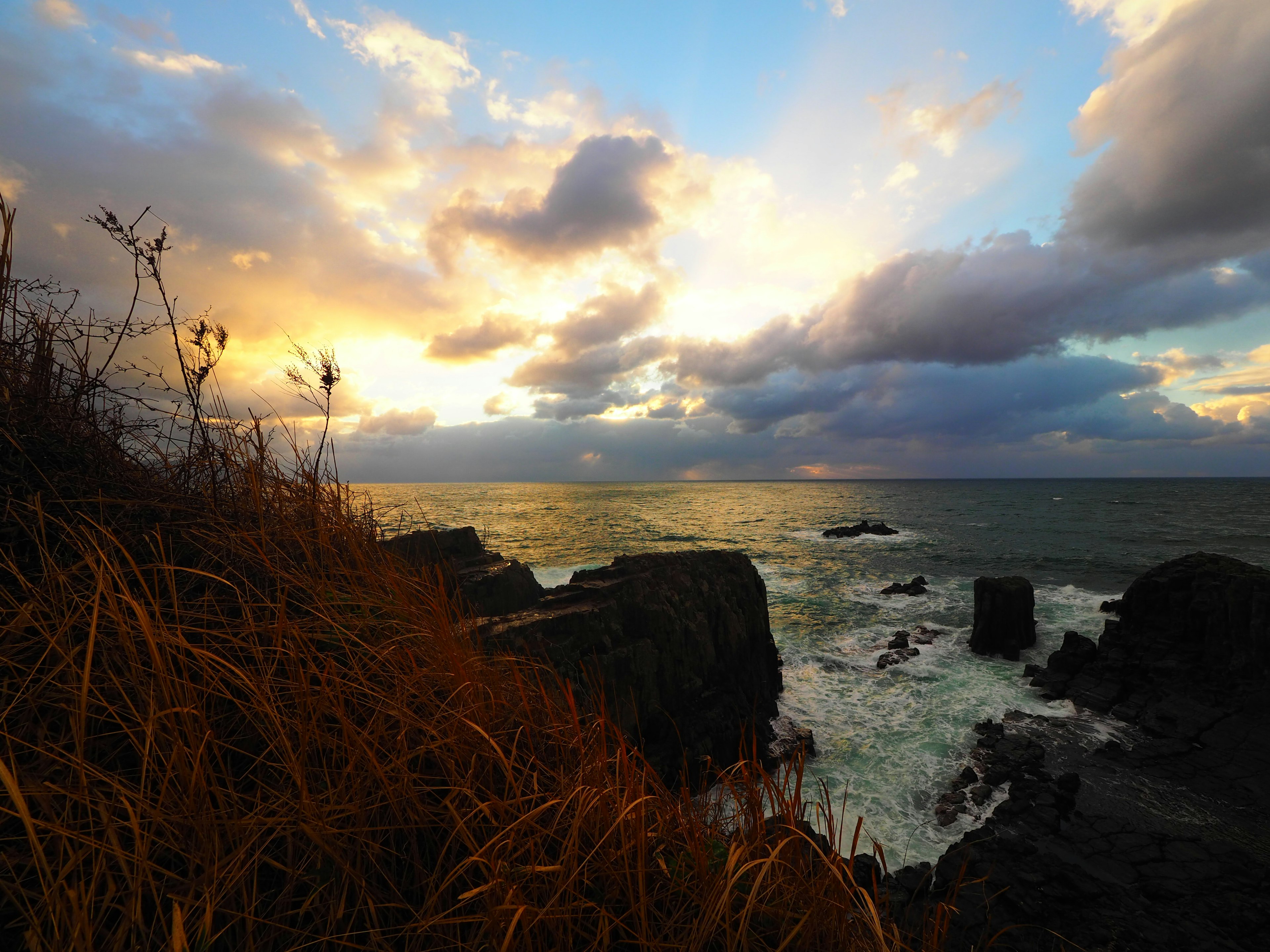 Pemandangan pantai saat matahari terbenam dengan awan dan ombak latar depan berbatu dengan rumput tinggi