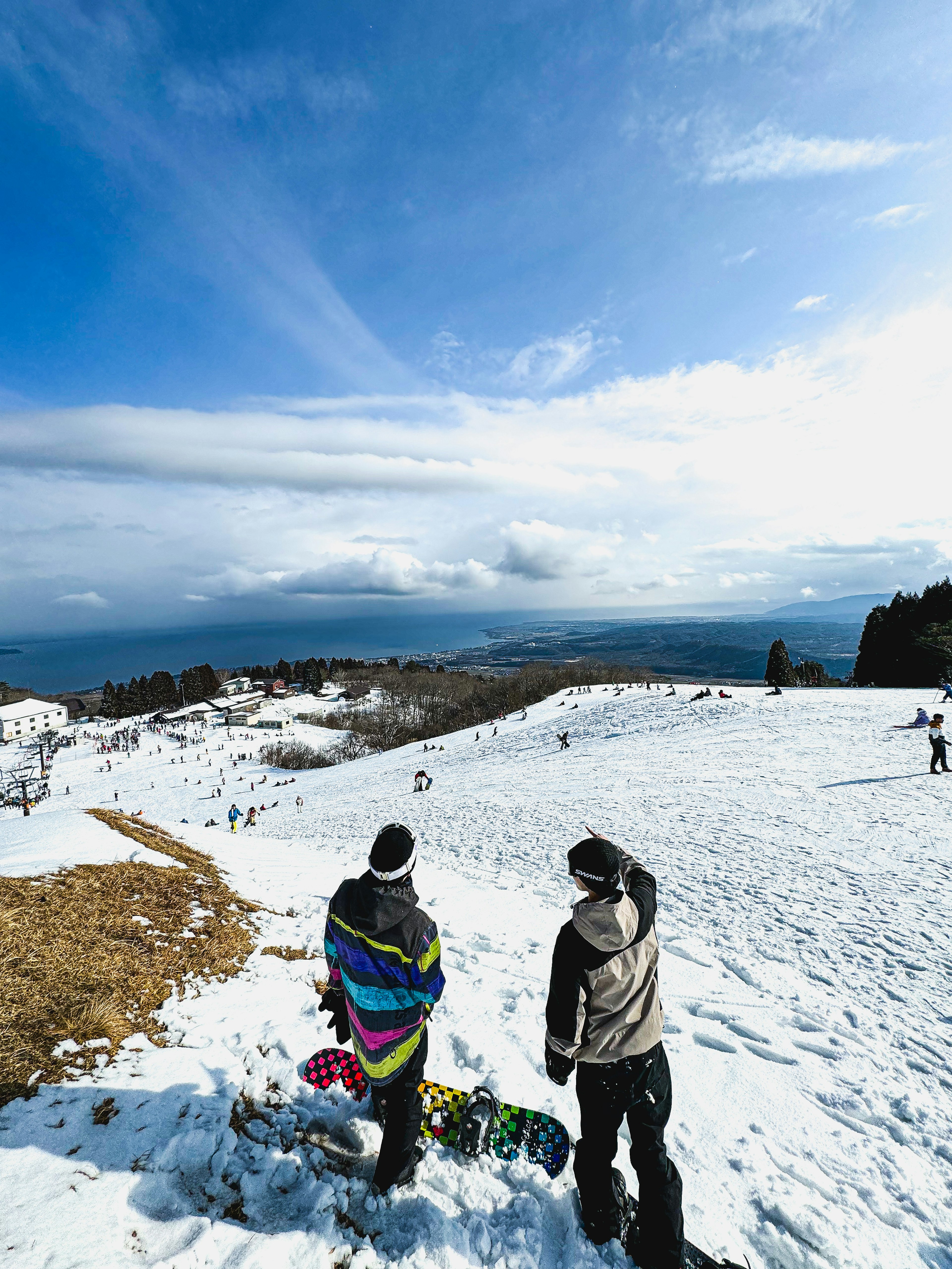 Two individuals with snowboards standing on a snowy slope overlooking a vast sky and mountains