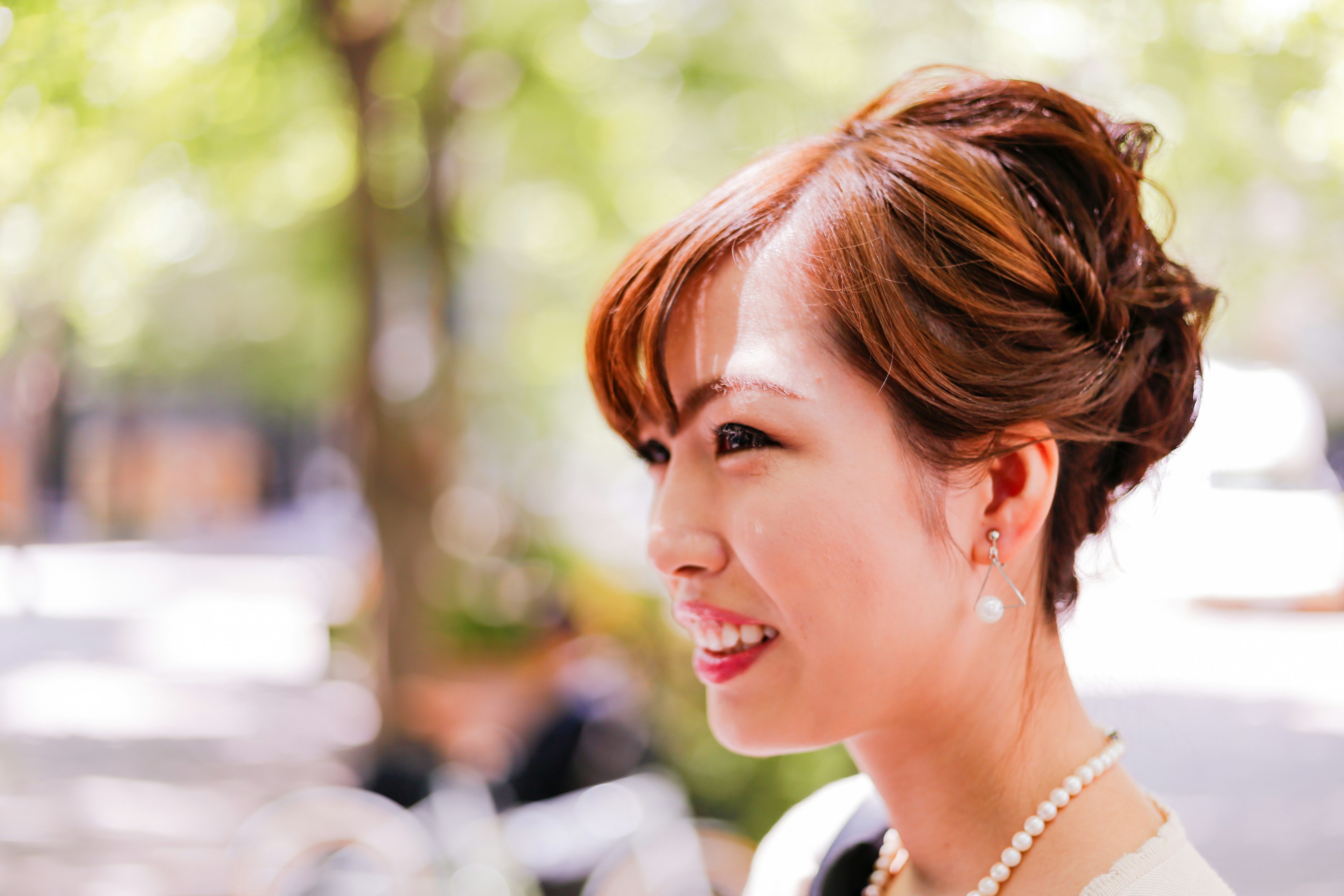 Profil d'une femme souriante dans un parc avec une coiffure en chignon portant des boucles d'oreilles en perles