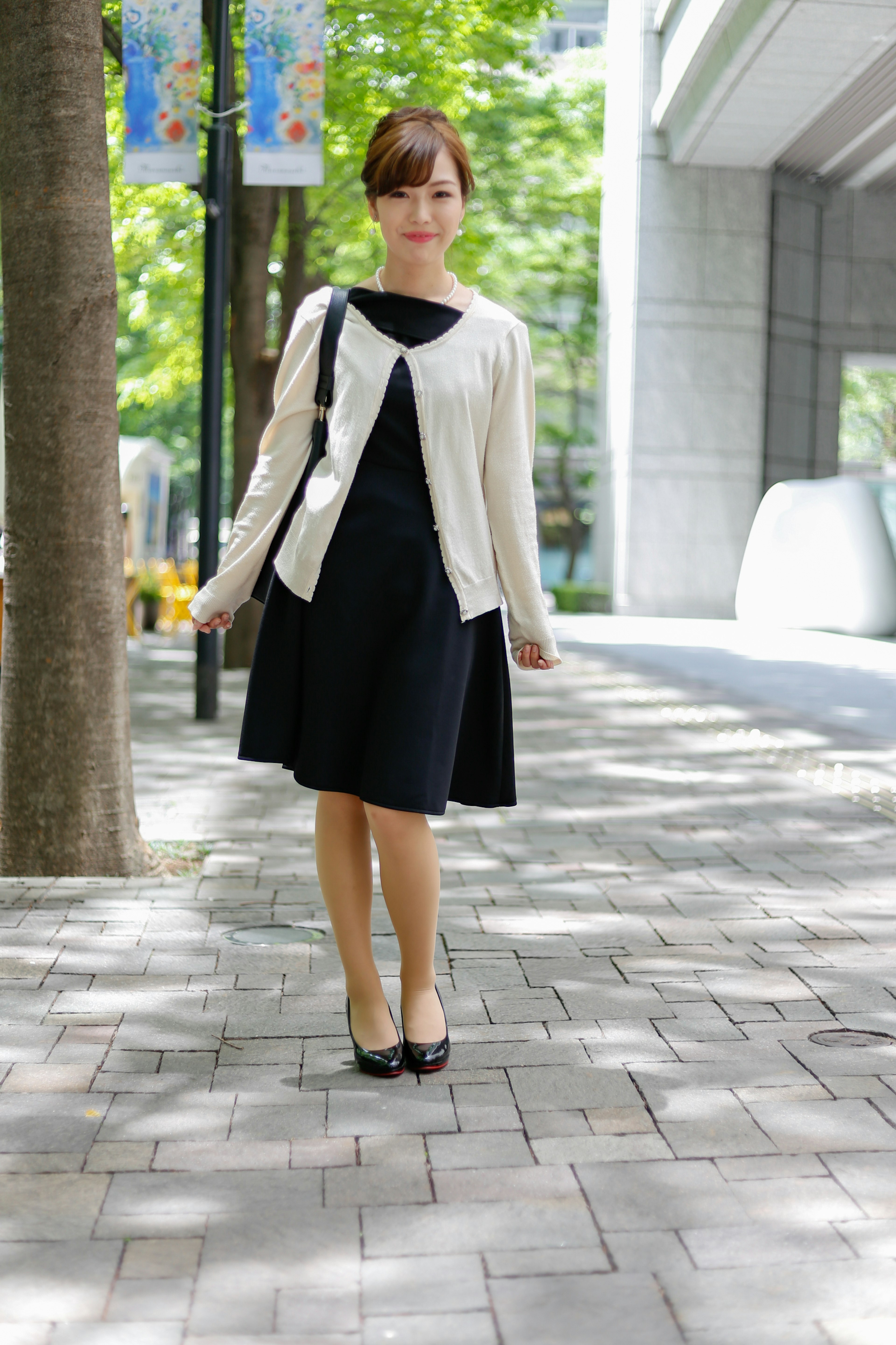 A woman in a black dress and white cardigan stands on a sidewalk